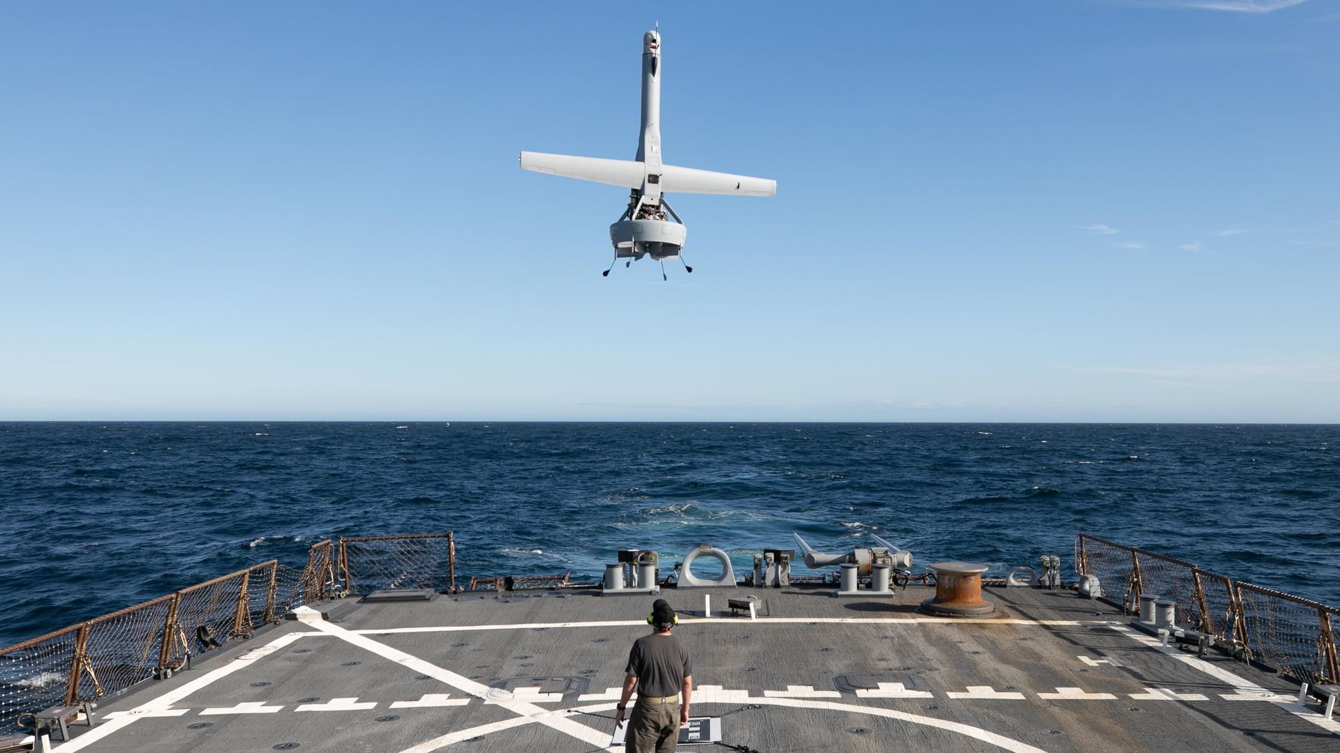 A VBAT drone, which looks like an inverted cross, hovers over the deck of a U.S. Navy ship at sea. A person stands underneath it.