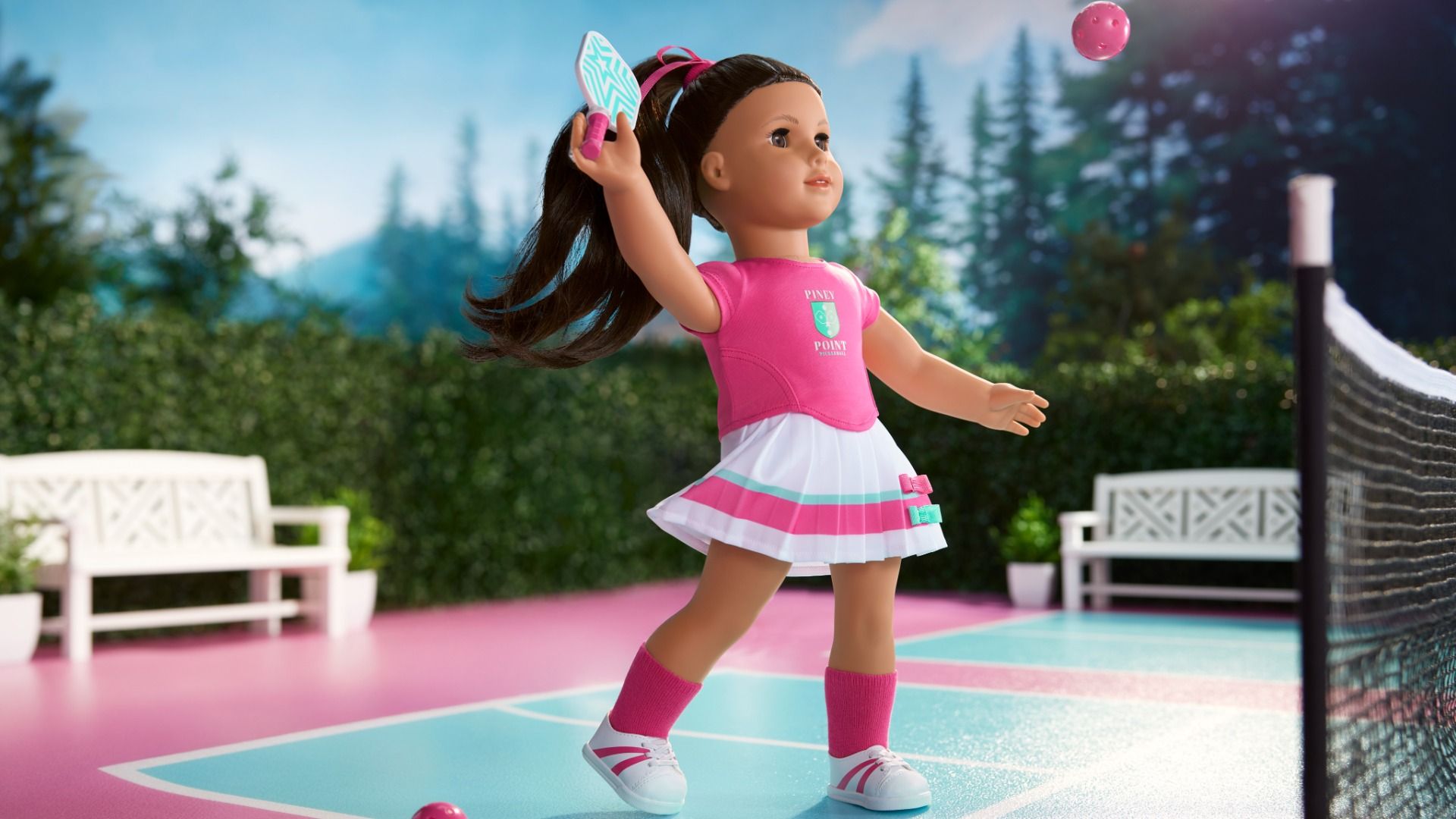 Doll with dark ponytail in pink and white outfit playing pickleball on a pink and blue court, holding paddle and hitting a pink ball, with green foliage and white benches in background.