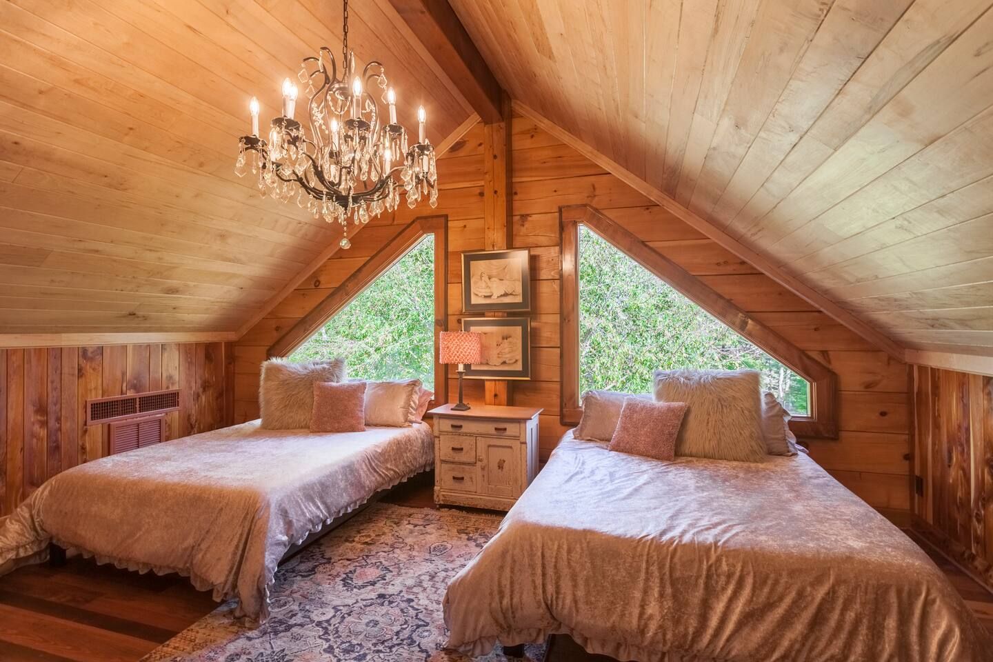 Cozy attic bedroom with wooden walls and ceiling, two beds with soft pink bedding and fluffy pillows, a chandelier, a vintage nightstand with a lamp, and triangular windows showing green trees.
