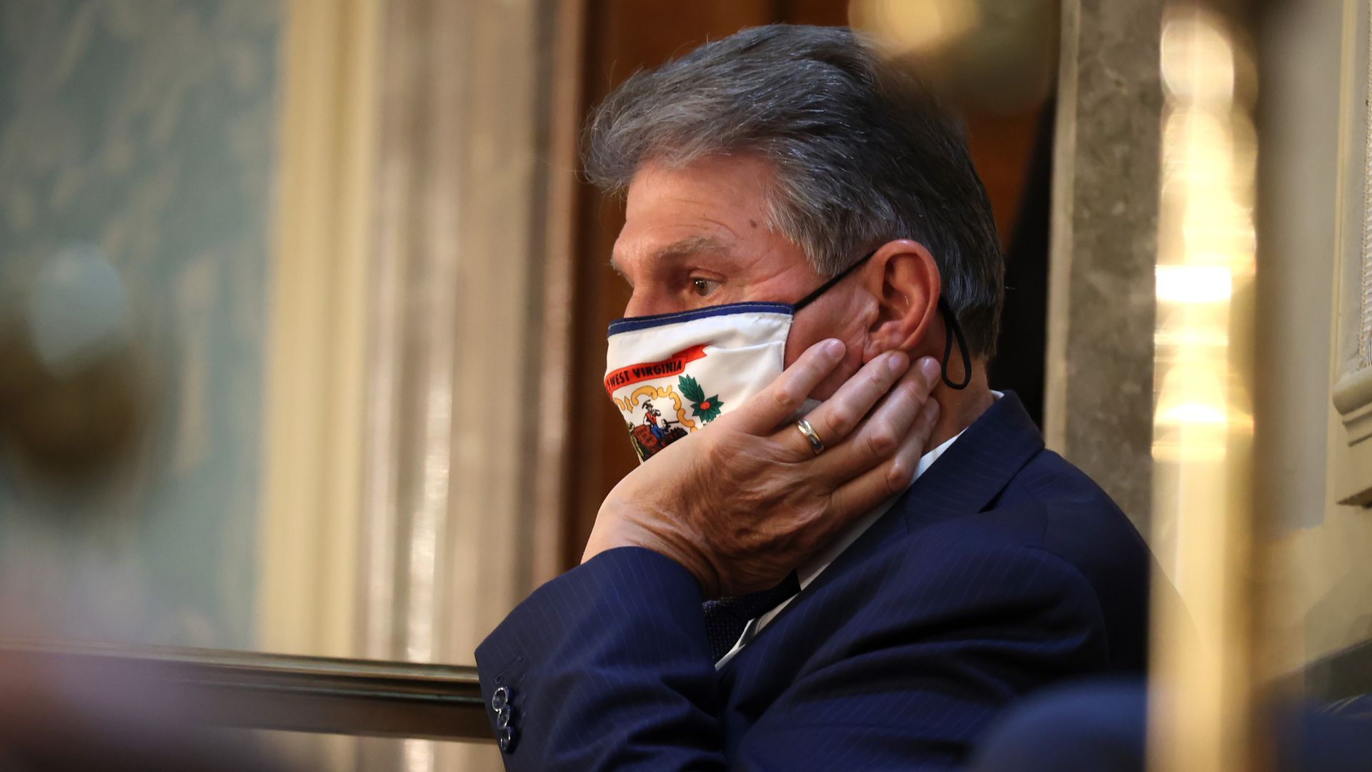 Joe Manchin sits with a hand resting against his face while wearing a face mask 