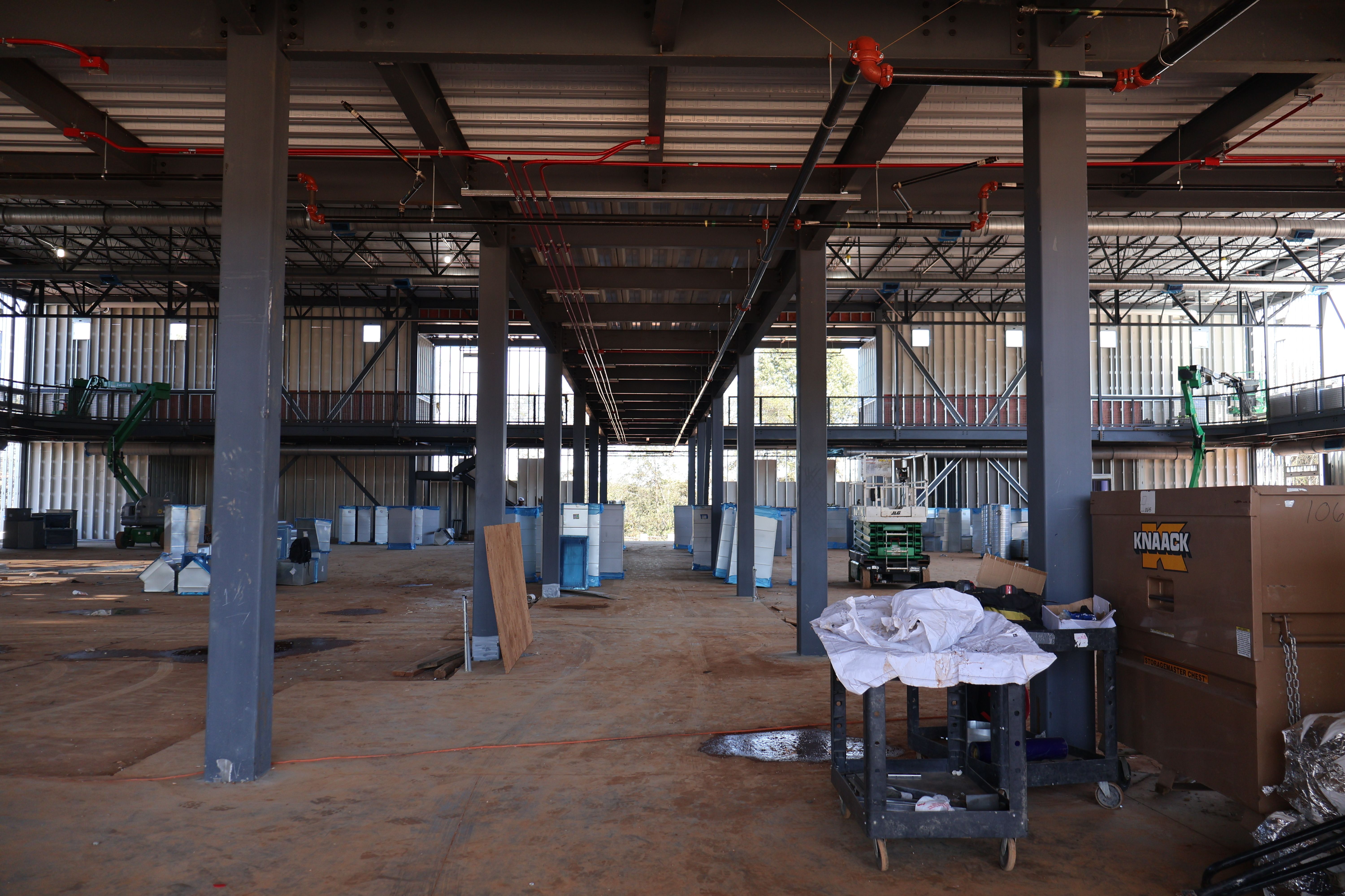 Interior of a spacious building under construction with metal beams, scattered equipment, machinery, and construction materials on a concrete floor.