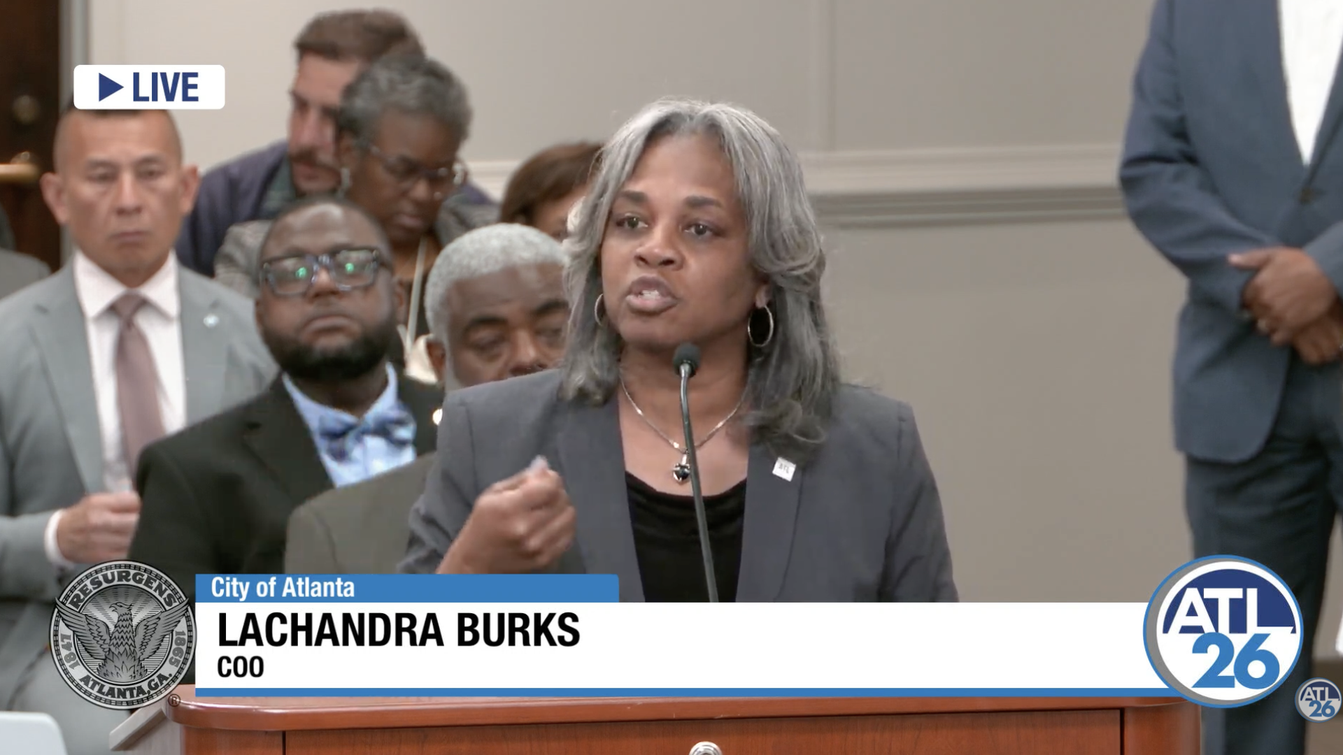 Gray-haired woman in a blazer speaks at a podium with a microphone; audience behind. A blue banner shows "City of Atlanta" and "LACHANDRA BURKS, COO"; a LIVE badge is visible.