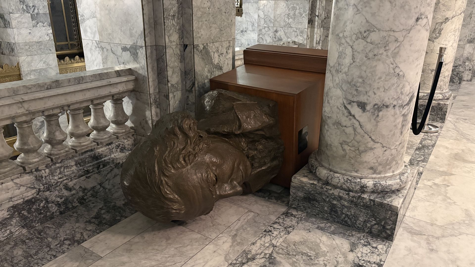 Large bronze bust sculpture of George Washington lying on its side on a marble floor, surrounded by marble columns and balustrades in an elegant indoor space.