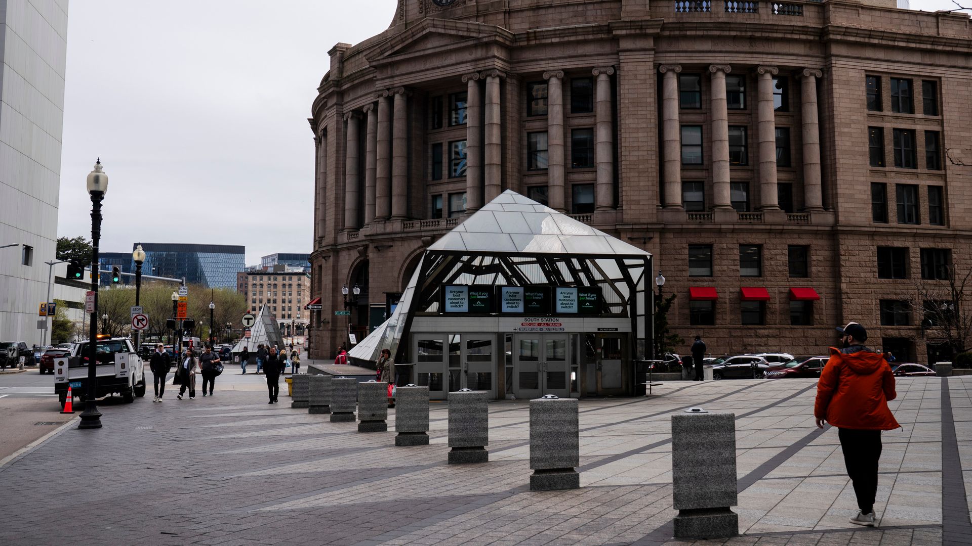 A photo showing South Station in Boston in the background and people walking near the glass-covered entrance to the Red Line trains below.