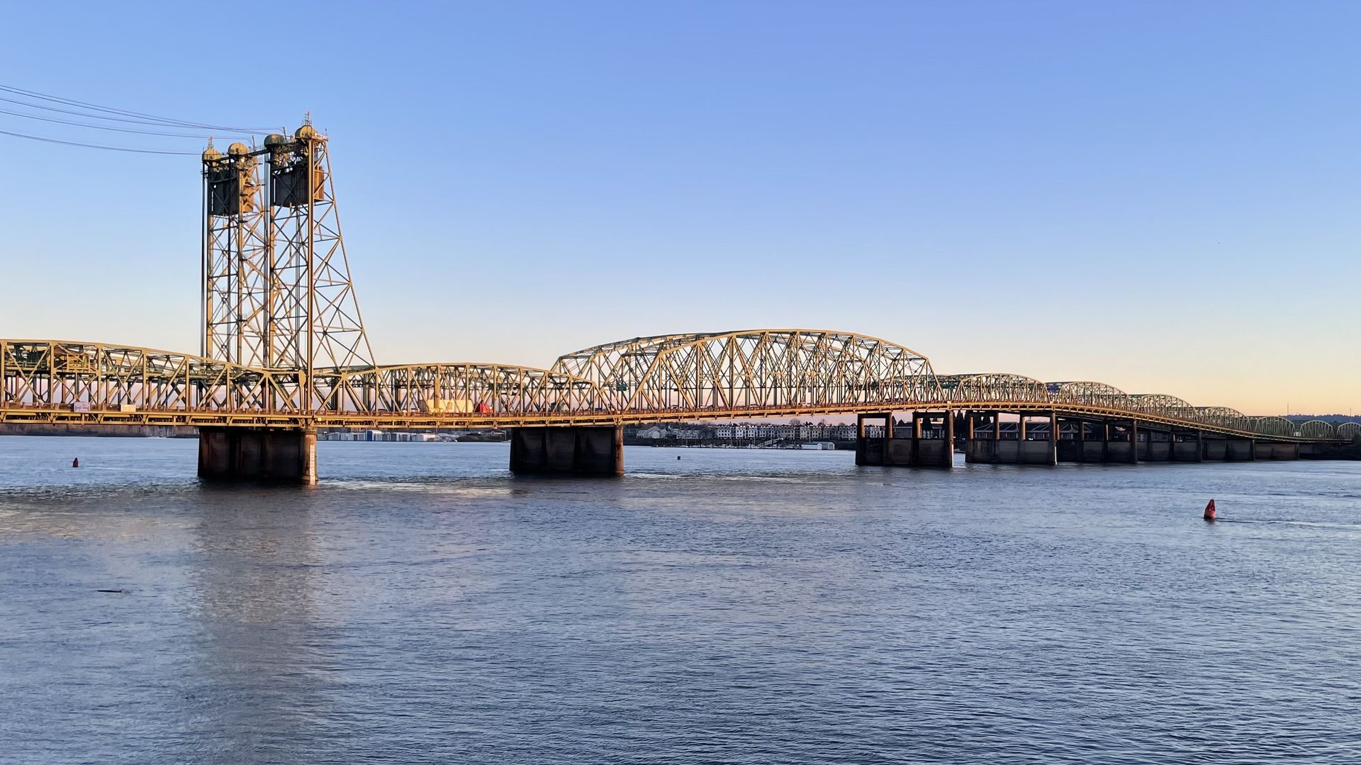 Wide river with a metal truss bridge stretching across, tall lift-span towers on the left, calm water reflecting the blue sky and orange sunset, distant shoreline with buildings.