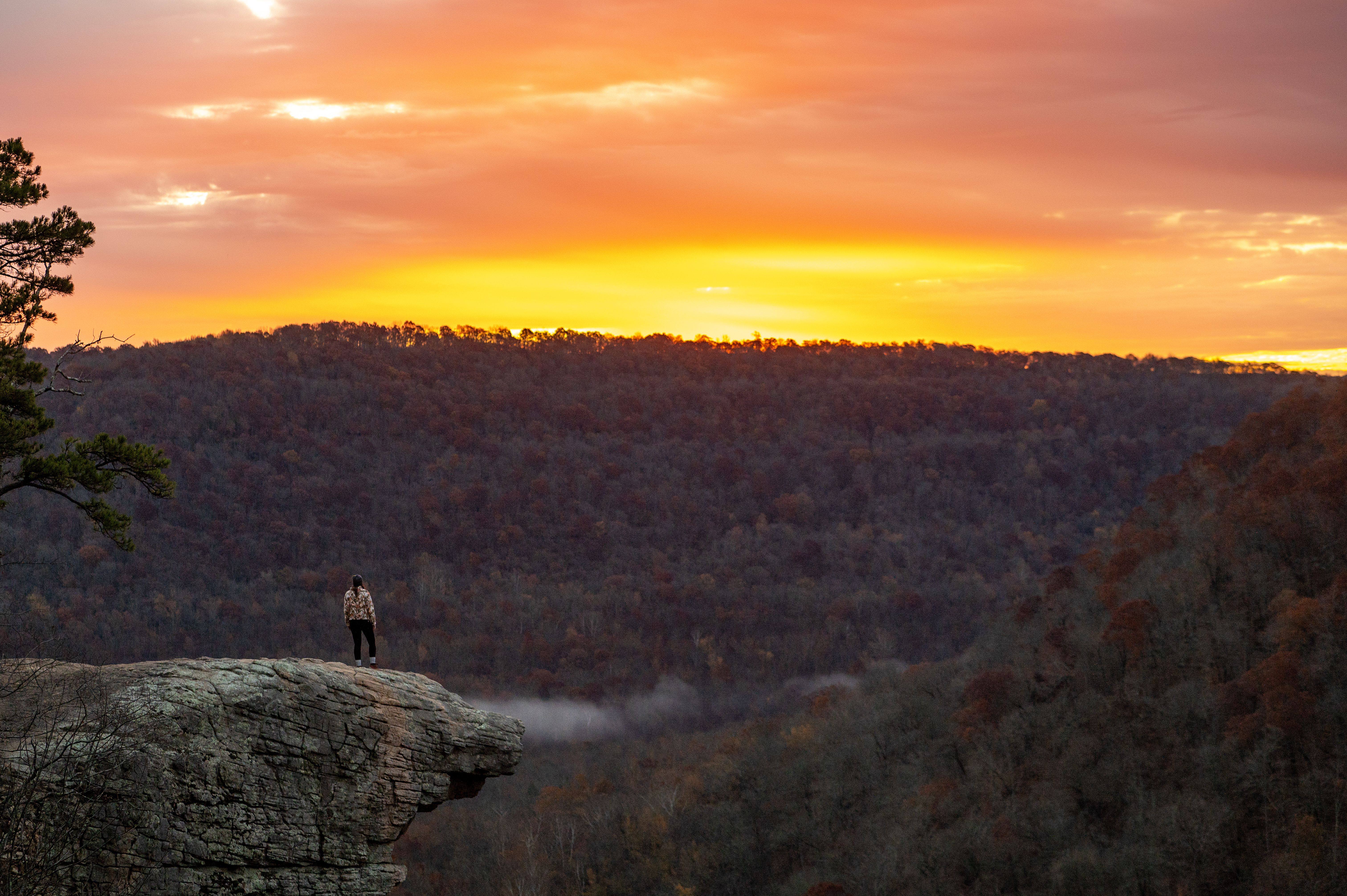 A person stands on a rocky cliff overlooking a forested valley at sunset, with a vivid orange and pink sky above.