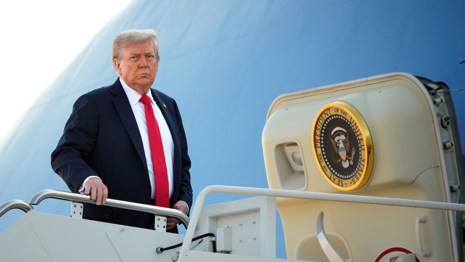Man in black suit and red tie standing on stairs beside Air Force One door with the presidential seal, against a light blue background.