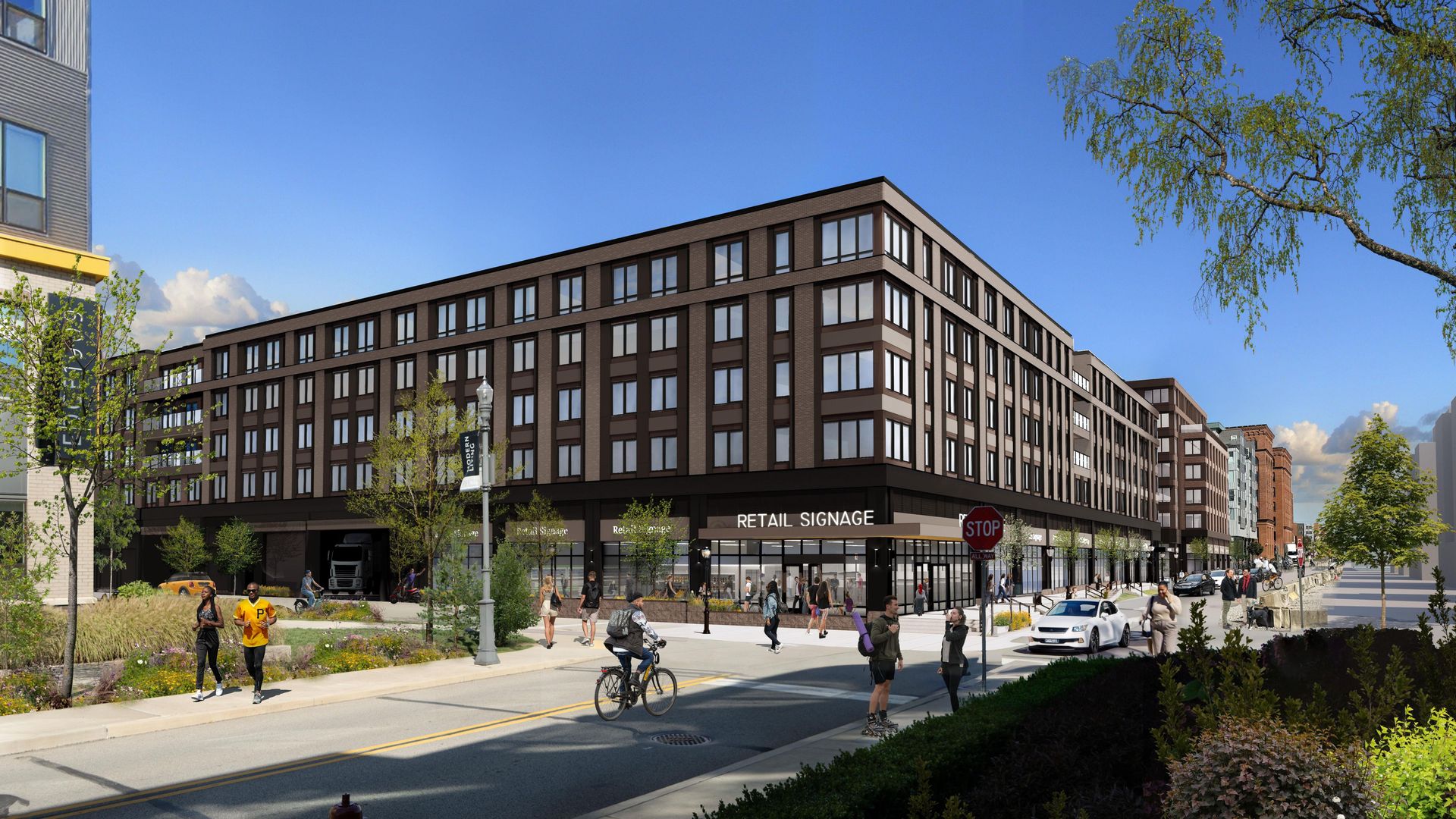 Modern five-story brown and beige building with retail signage at street level, people walking, jogging, biking, and cars on a sunny day with blue sky and greenery around.