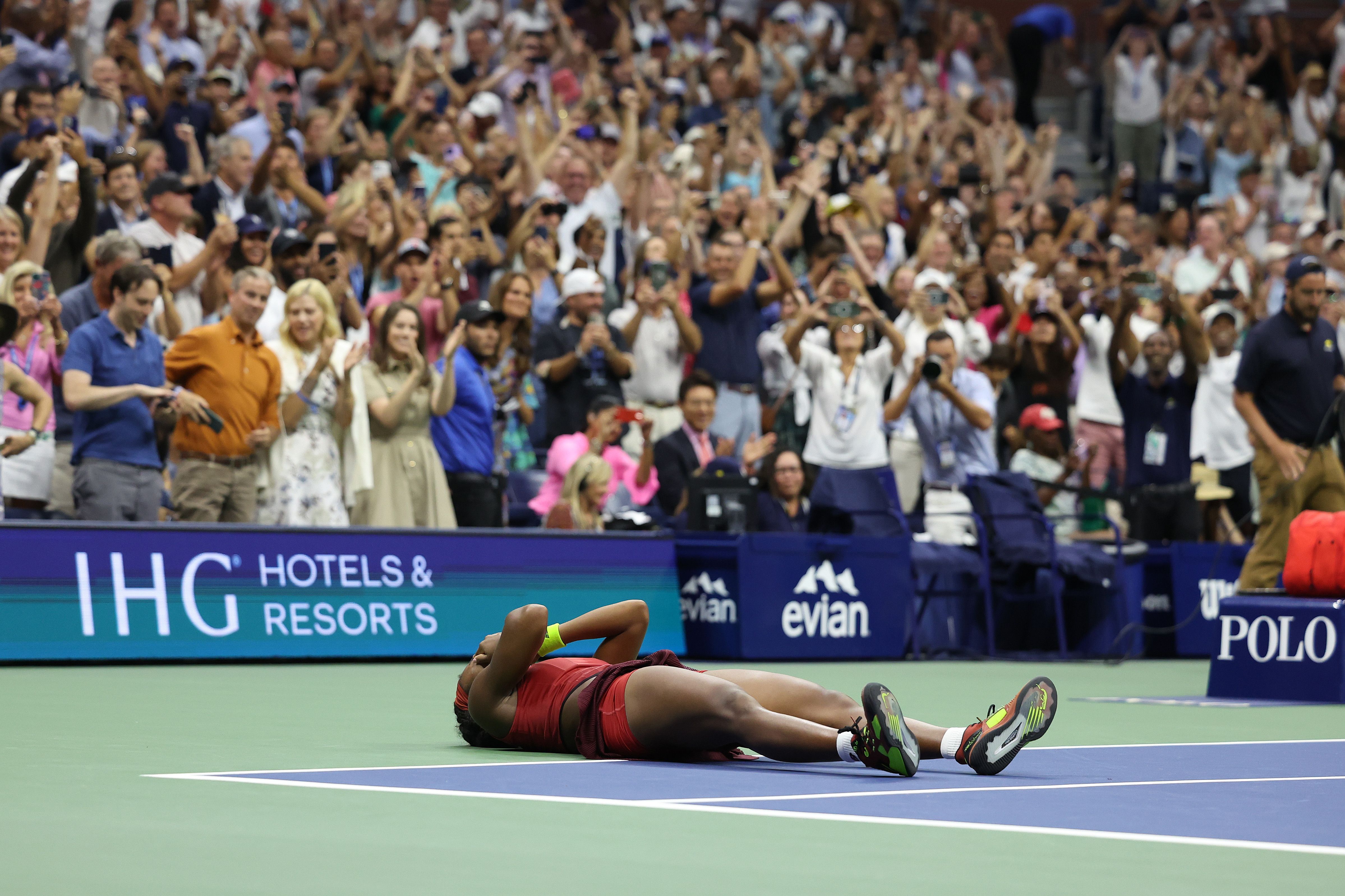 An image of Coco Gauff laying down on a tennis court with her hands over her face, people are cheering in the background. 