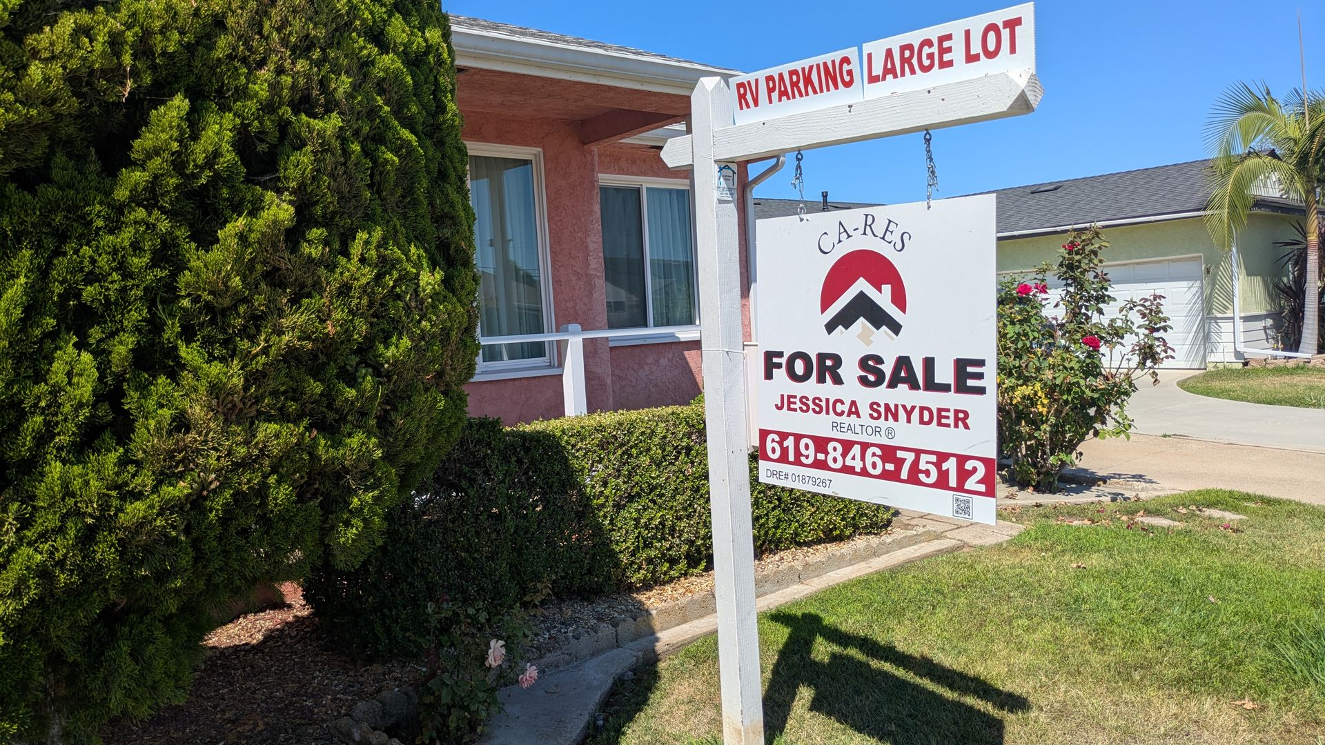 Sunny suburban scene with a pink stucco house, tall green hedge, and a white real estate sign reading 'RV PARKING LARGE LOT' above a 'FOR SALE' board; blue sky and a palm tree visible.