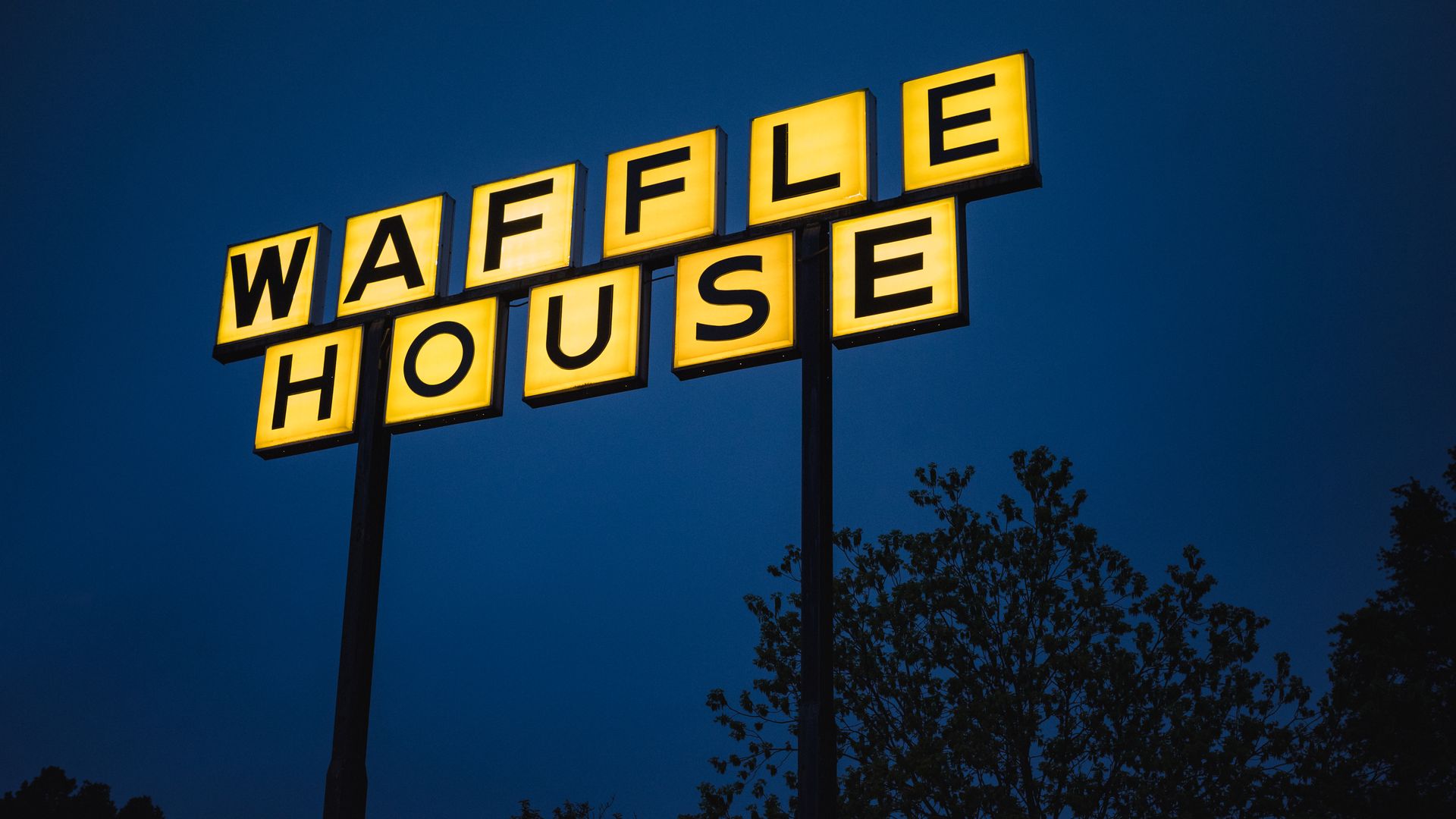 Two yellow, illuminated sign blocks spell "WAFFLE HOUSE" on two posts, set against a dark blue evening sky with silhouetted trees.