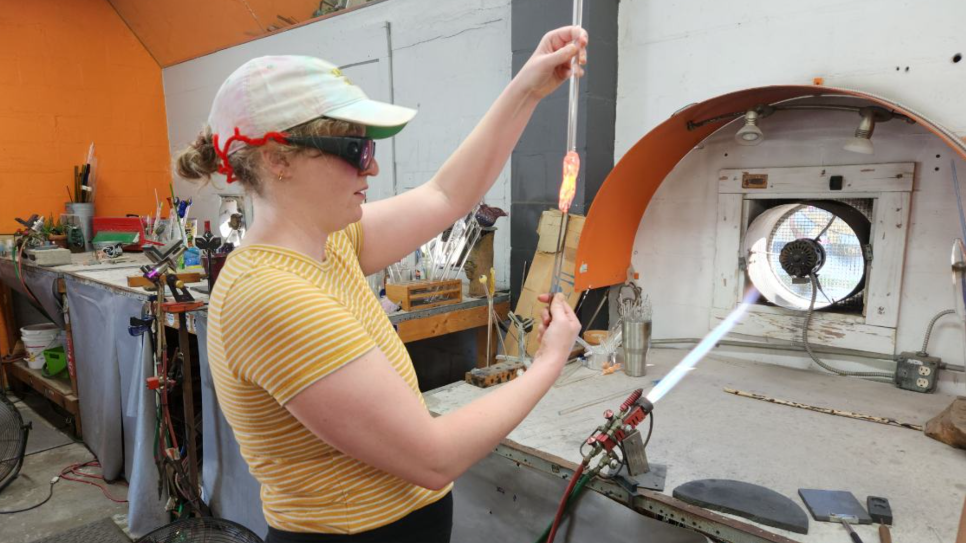 A woman in a yellow top handling molten glass with a pair of tweezers.