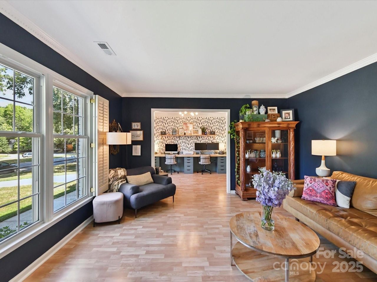 Living room with dark blue walls, large windows, gray and brown leather sofas, wooden coffee table with purple flowers, wooden cabinet, and visible office space with patterned wallpaper in background.