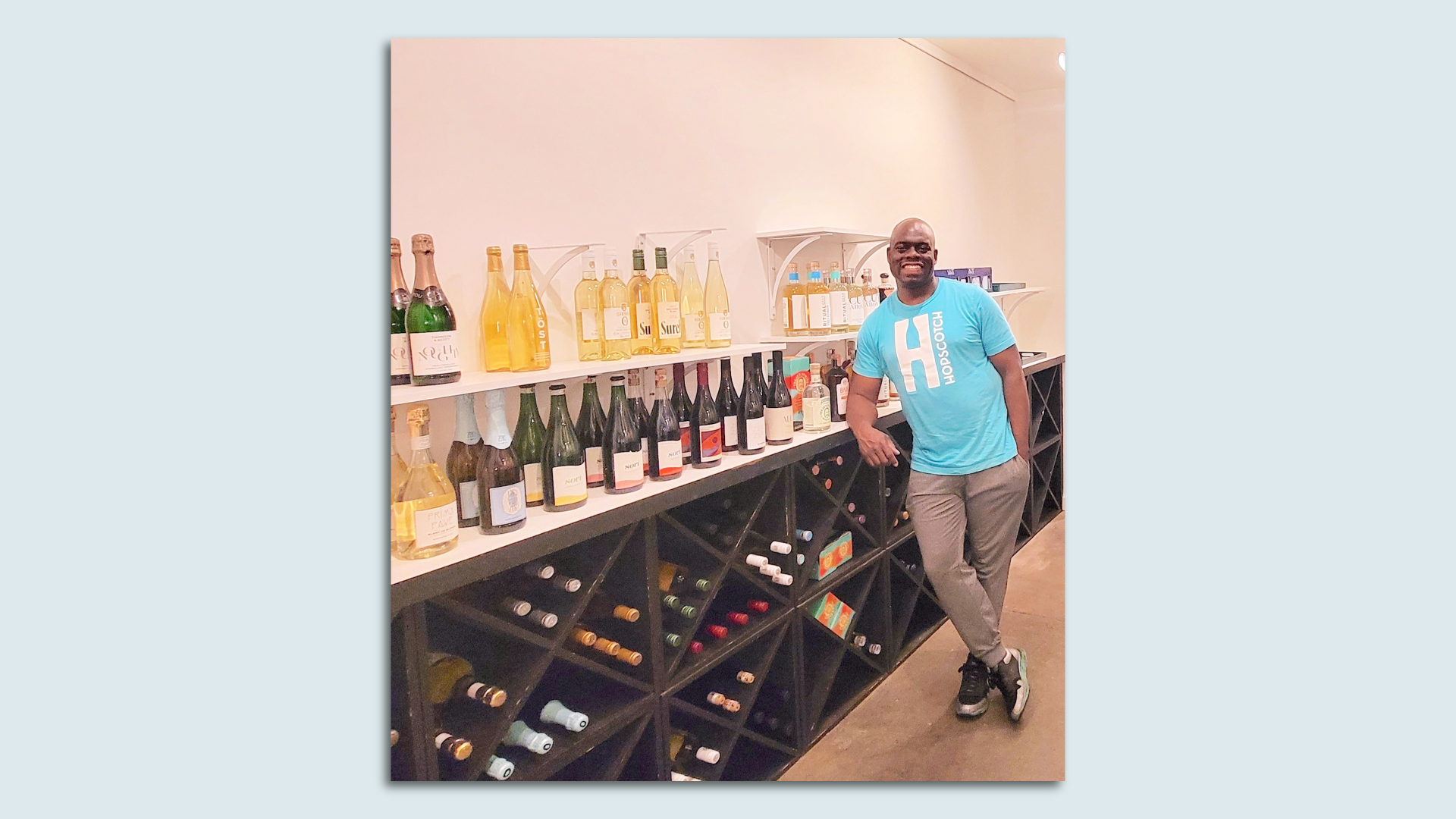 A man in a blue t-shirt stands in front of a wine rack filled with non-alcoholic wines 