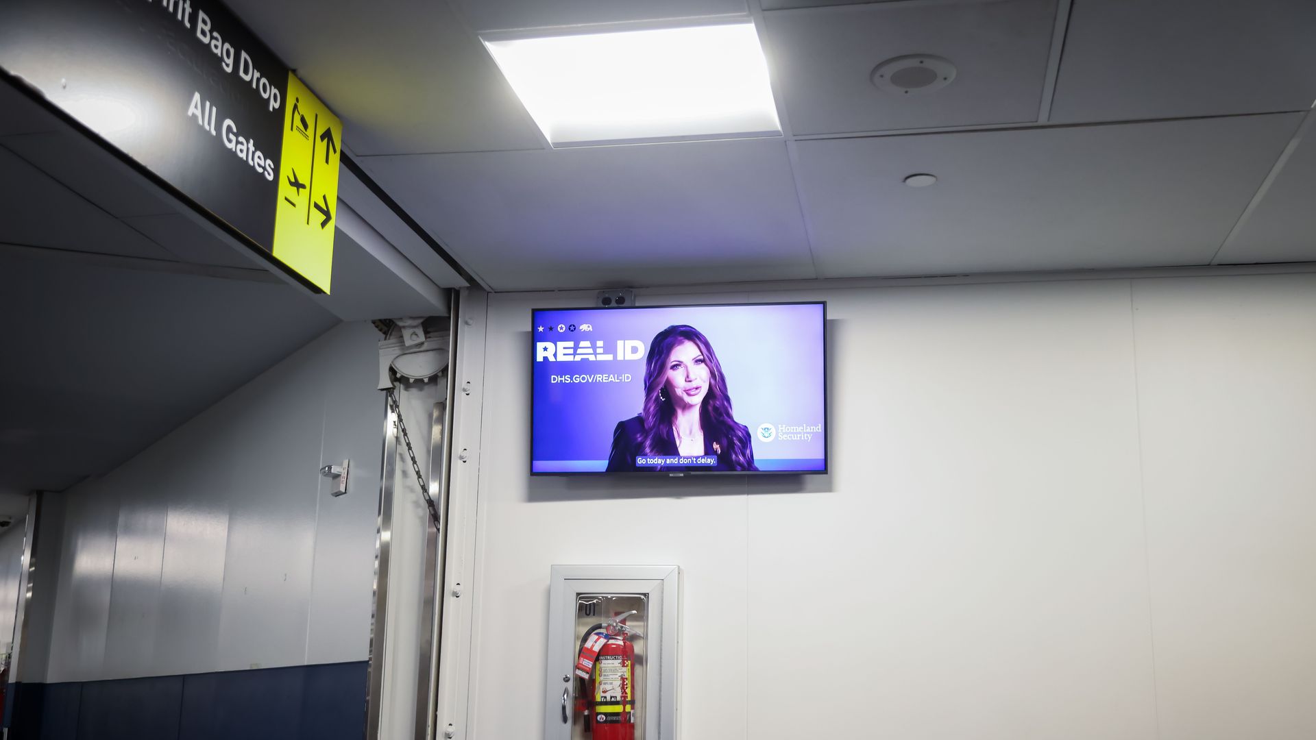 DHS Secretary Kristi Noem speaks on a TV screen at an airport in front of the words "REAL ID."