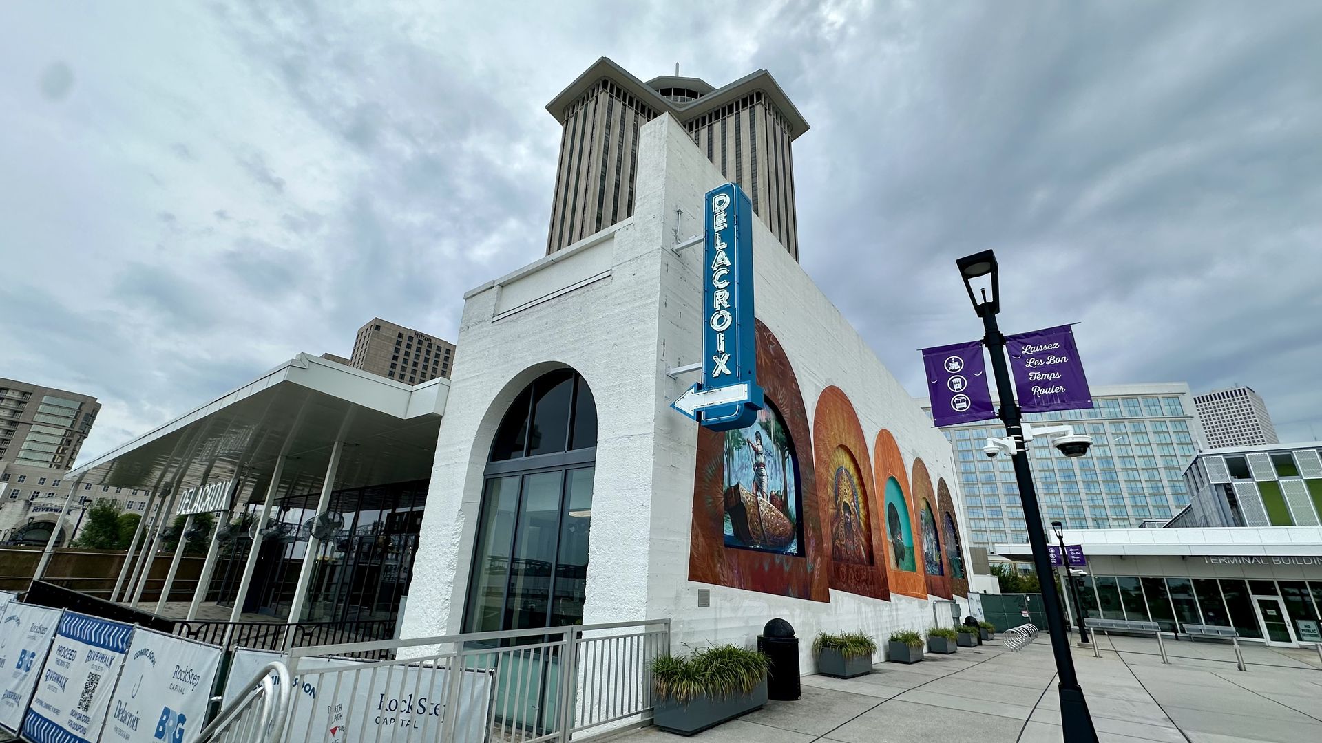 White building with blue "DELACROIX" vertical neon sign and colorful murals under cloudy sky; modern buildings and purple banners saying "Laissez Les Bon Temps Rouler" visible in urban plaza.