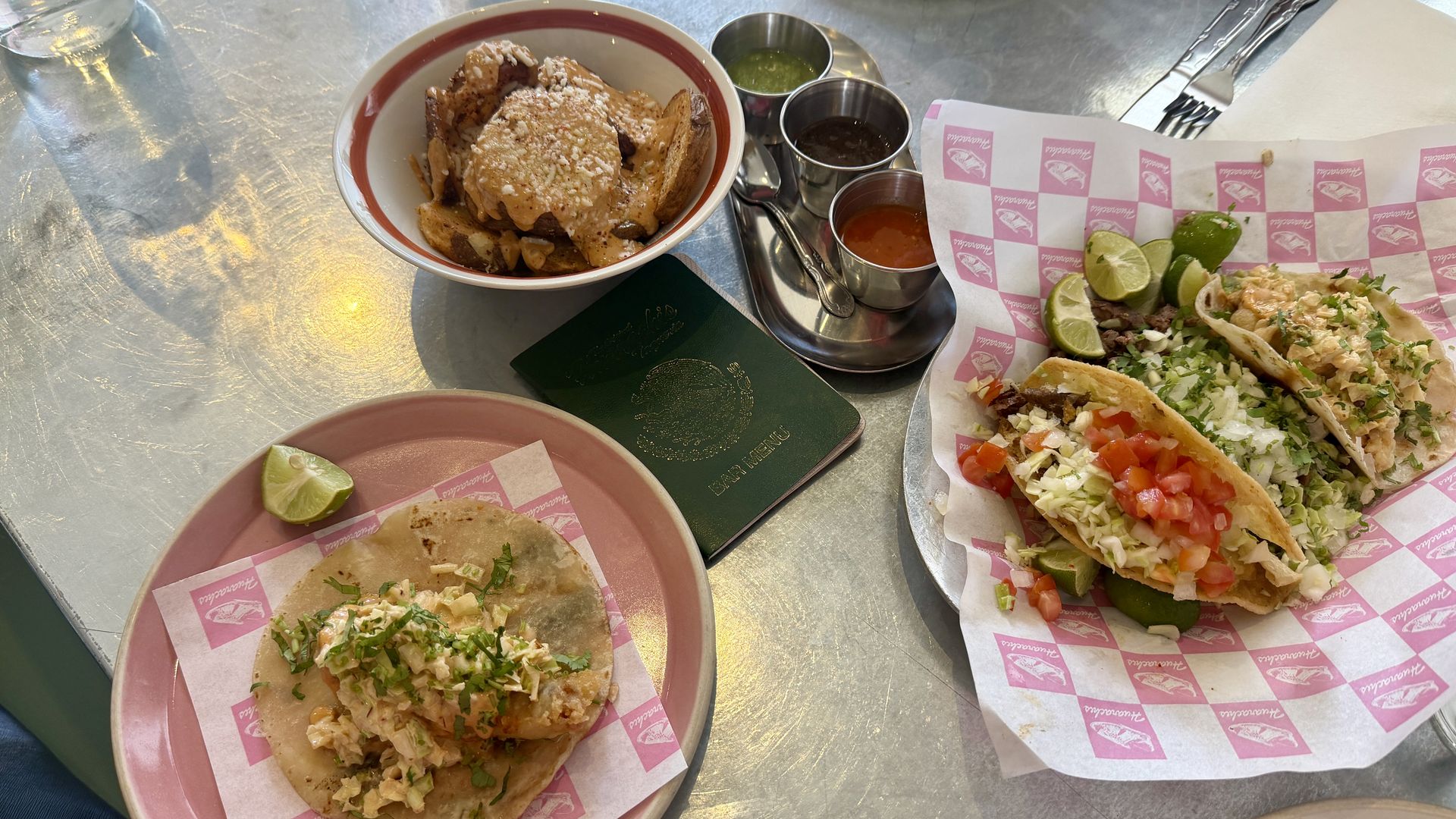 Top-down view of a metal table with tacos and toppings: a pink plate taco with cilantro, a tray of two tacos, a bowl of potatoes, lime wedges, and metal sauce cups; a green menu nearby.