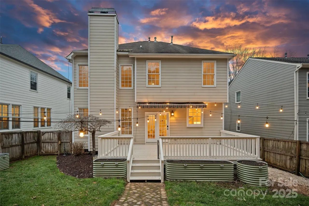 Back view of a beige two-story house with lit windows and string lights over a white wooden deck at sunset with purple and orange clouds, fenced backyard, and green grass.