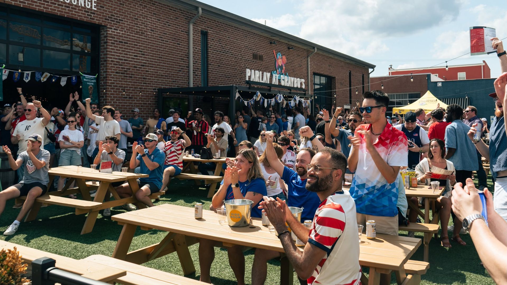 Crowd gathered outside a brick bar and lounge, seated at picnic tables and cheering. The building shows a "BAR AND LOUNGE" sign, string lights, and a blue sky with scattered clouds.