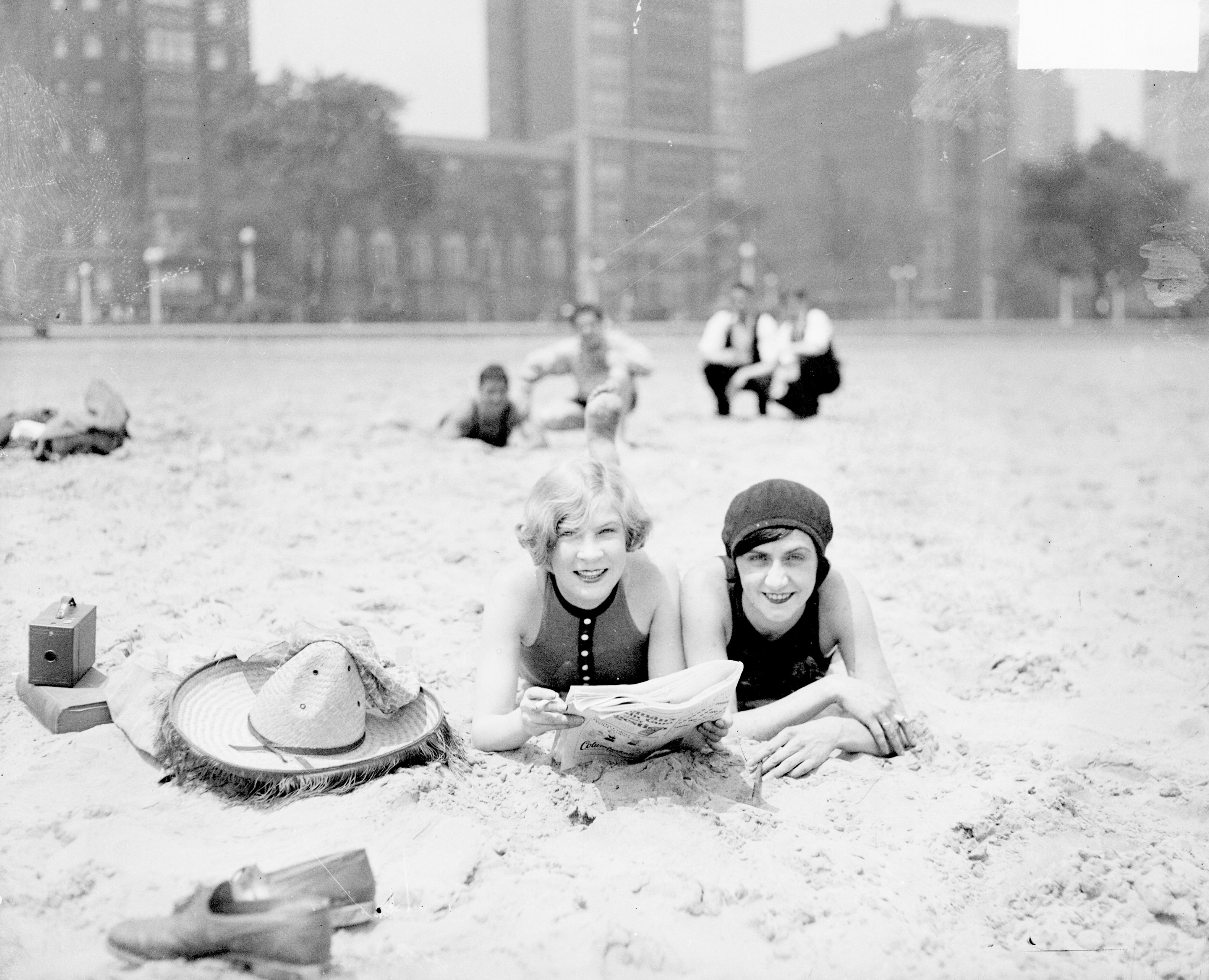 Black-and-white photo of two women in swimsuits lying on a sandy beach, smiling at the camera while reading a newspaper; hats and a box sit nearby, with a blurred cityscape in the background.