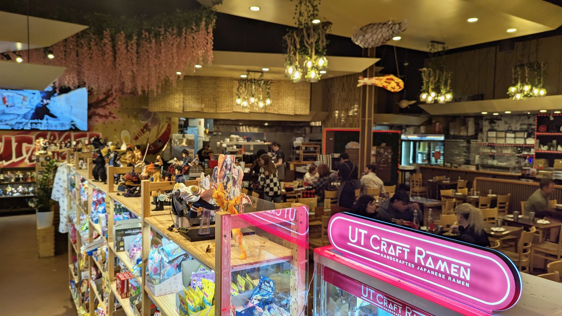 A pink neon sign reads "UT Craft Ramen" with shelves of toys nearby and a restaurant dining room behind it. 