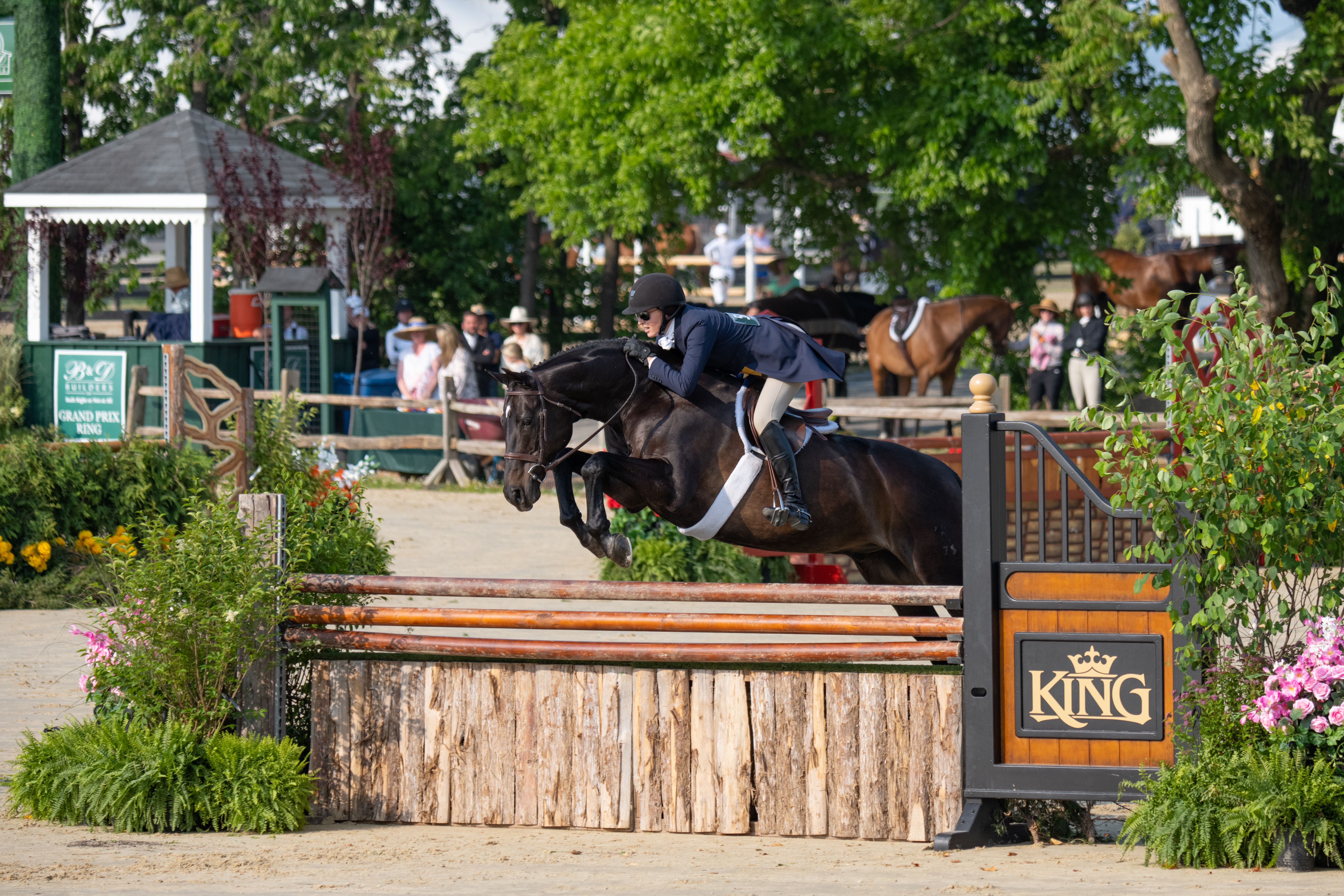 Rider in a navy jacket and helmet jumps a dark horse over a wooden orange-rail obstacle at an outdoor show, while spectators watch from under leafy trees by a pavilion.