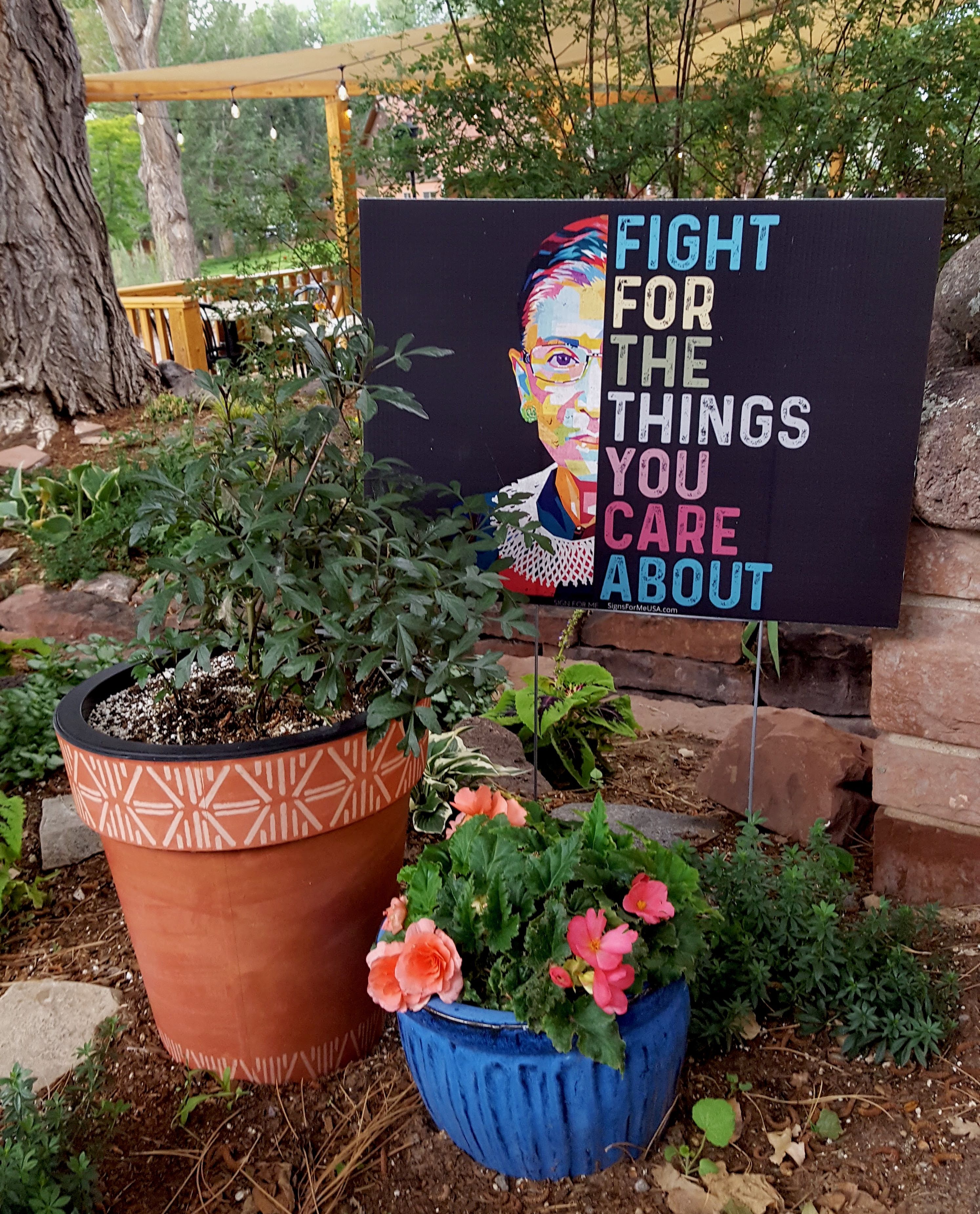A yard sign showing a photo of Ruth Bader Ginsberg next to the words "FIGHT FOR THE THINGS YOU CARE ABOUT" with a restaurant patio in the background and potted flowers in front of it.