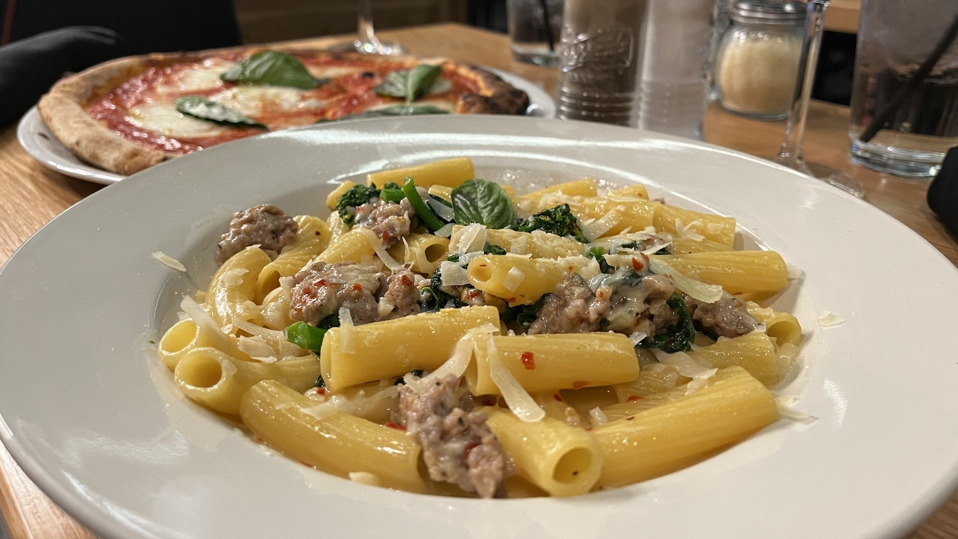 A bowl of pasta with a pizza in the background. 