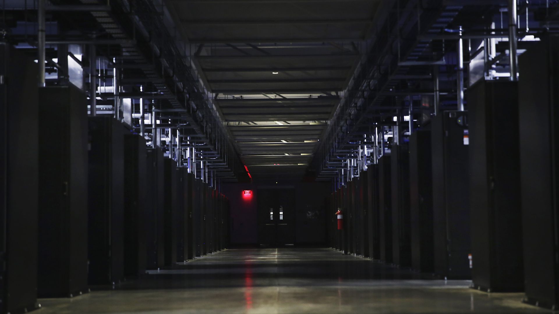 Dark industrial corridor with rows of pillars and overhead ducts, metal racks along both sides. The perspective converges to a distant lit exit; cool blue lighting with a red exit sign.