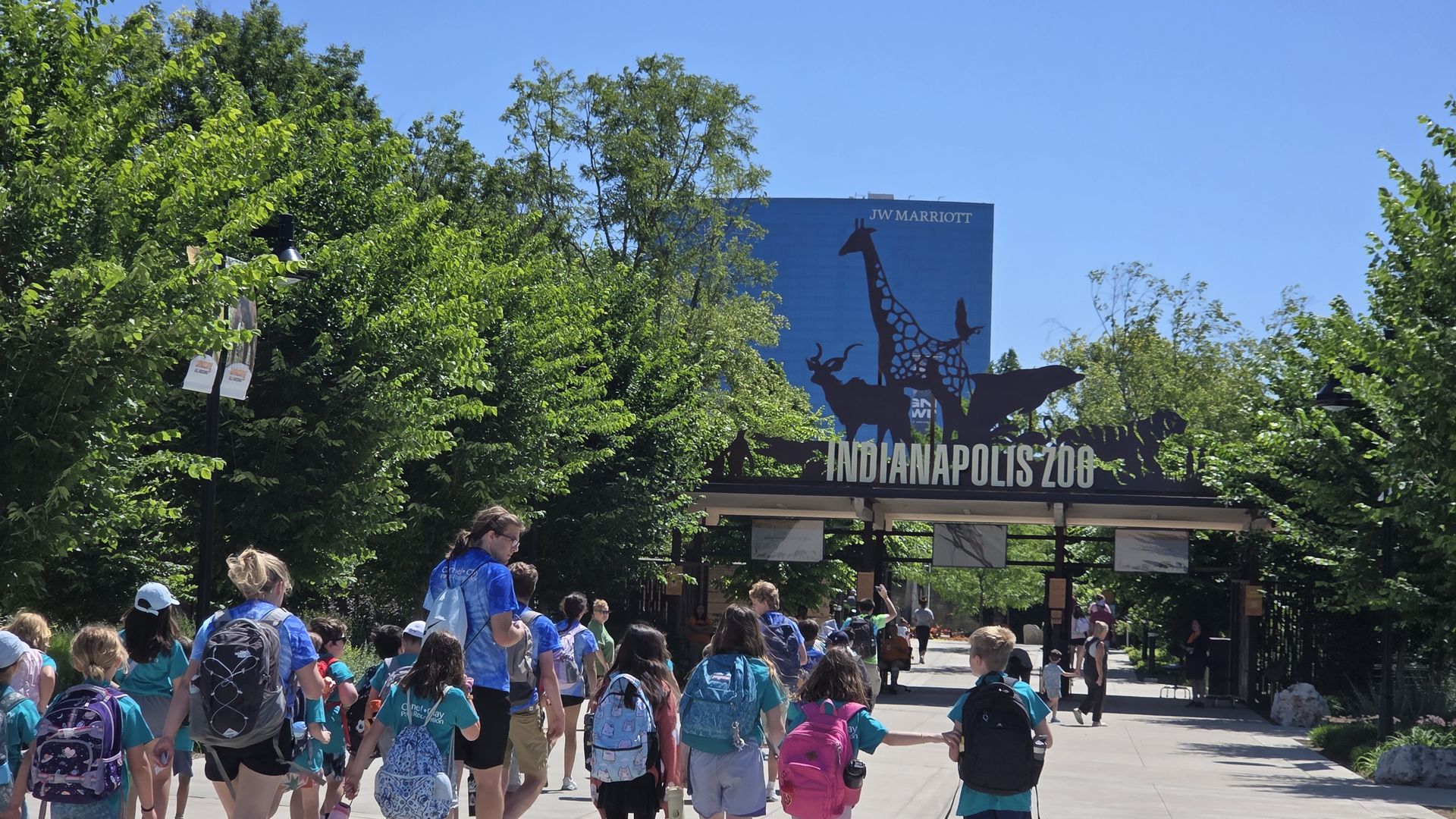 Sunny day at the Indianapolis Zoo entrance; a blue JW Marriott sign with a giraffe silhouette. Crowds of kids with backpacks walk past lush green trees toward the entrance.