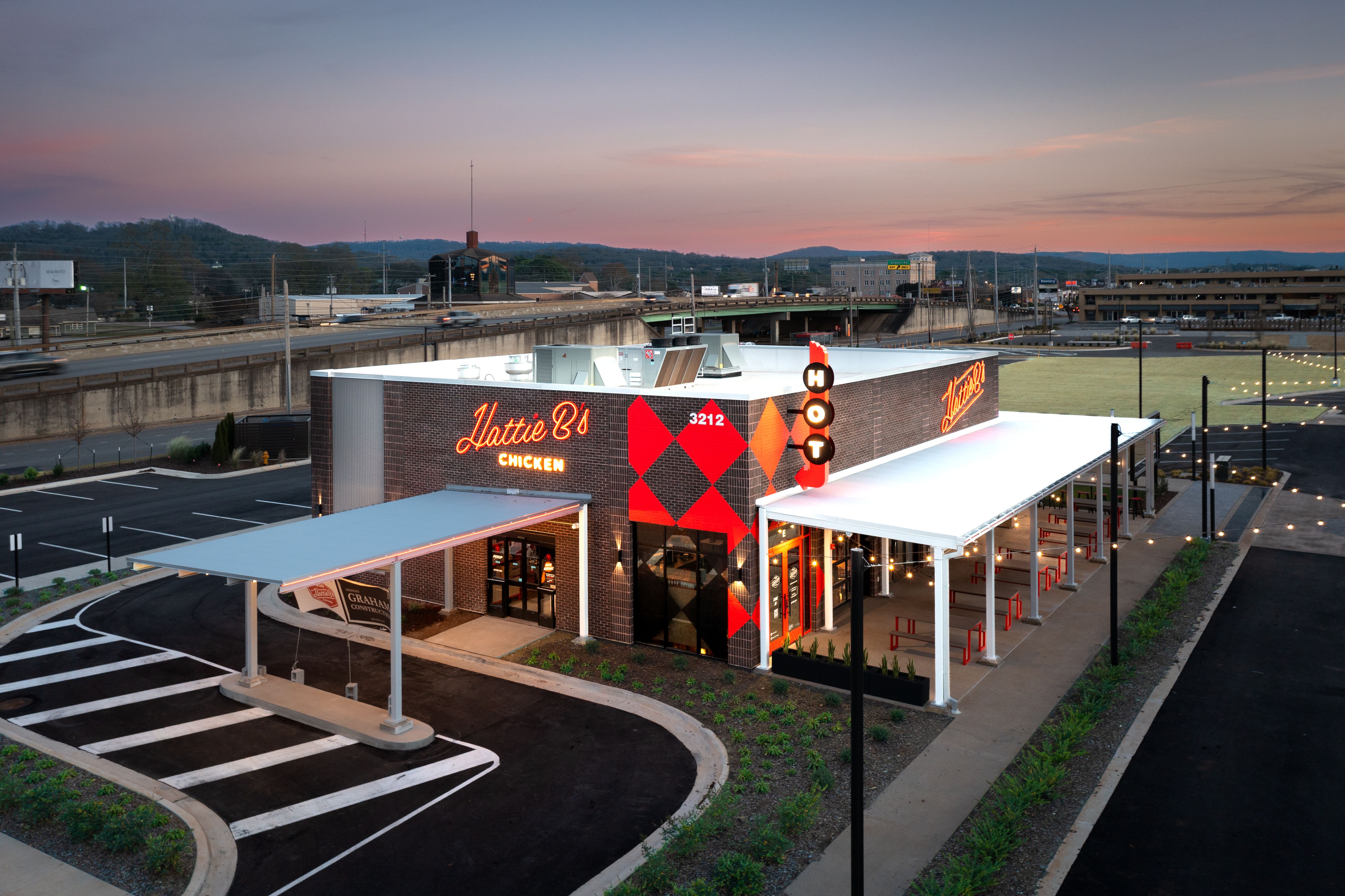 Aerial view of Hattie B's Chicken restaurant at dusk with bright orange neon signage, red and black diamond pattern on the brick building, outdoor covered seating with red stools, and nearby highway.