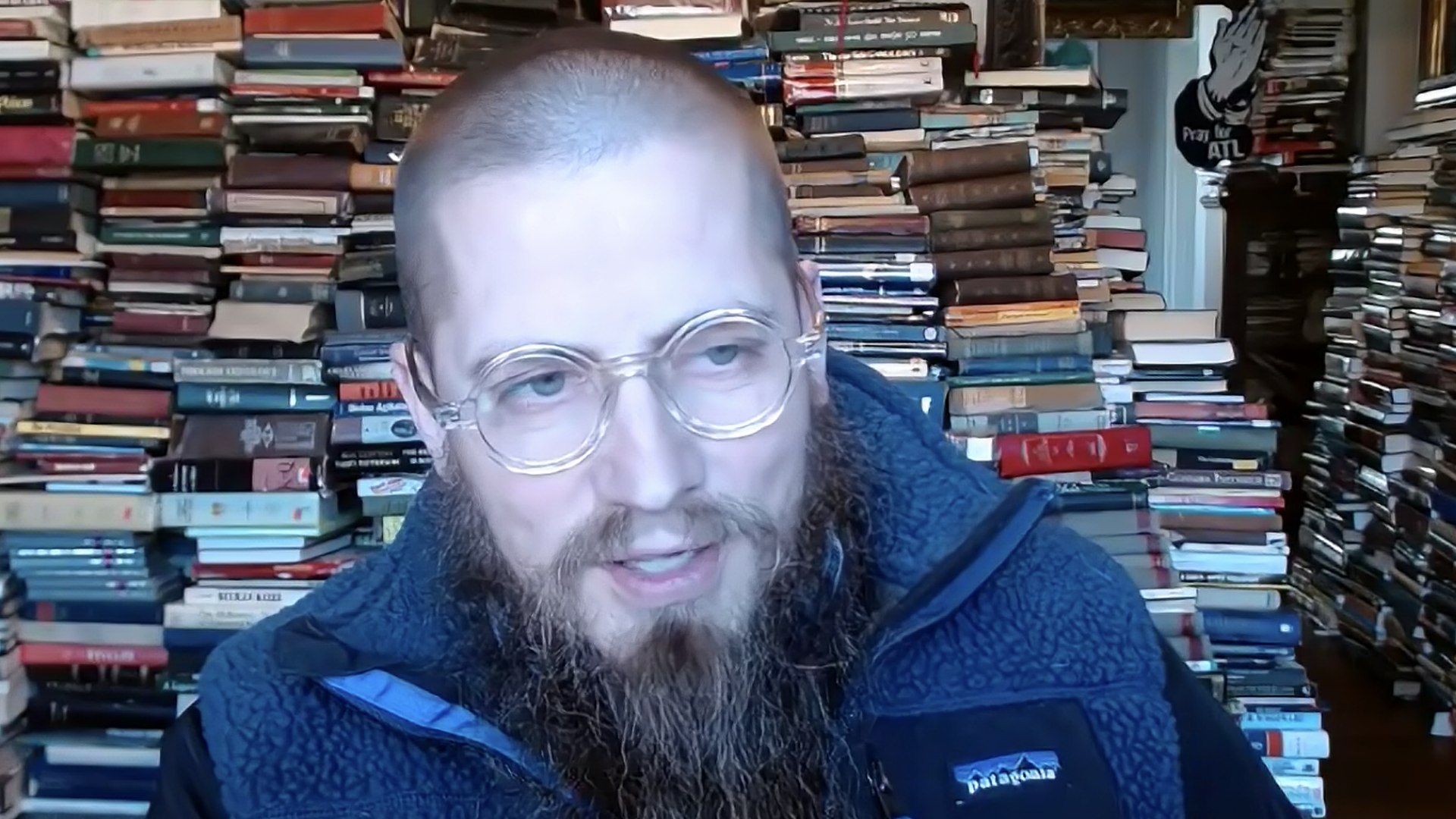 A screenshot of a man with a beard on a Zoom call with man books piled behind him. 