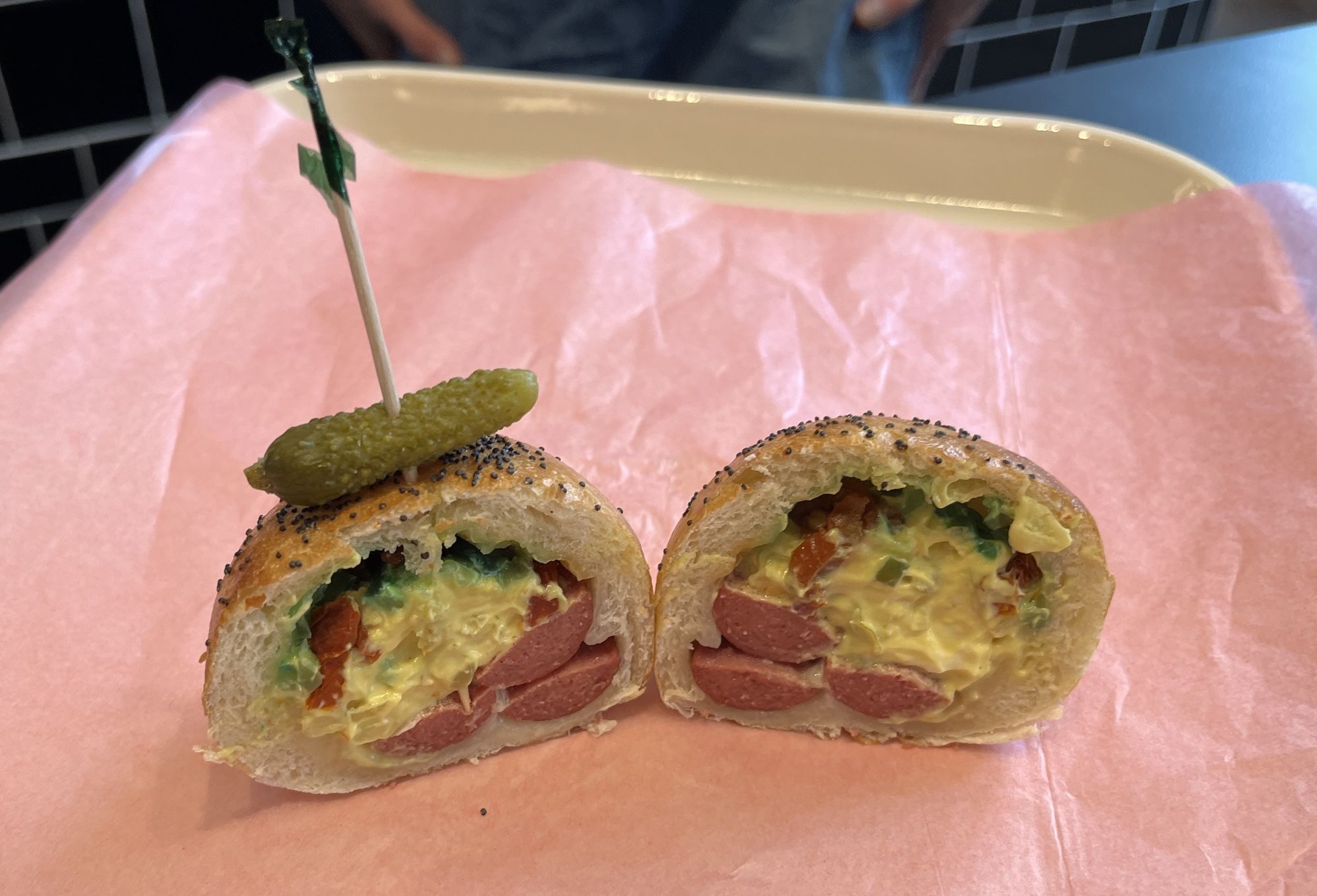 Poppyseed bun cut open with creamy mustard, slices of hot dogs, relish, and a pickle on top.