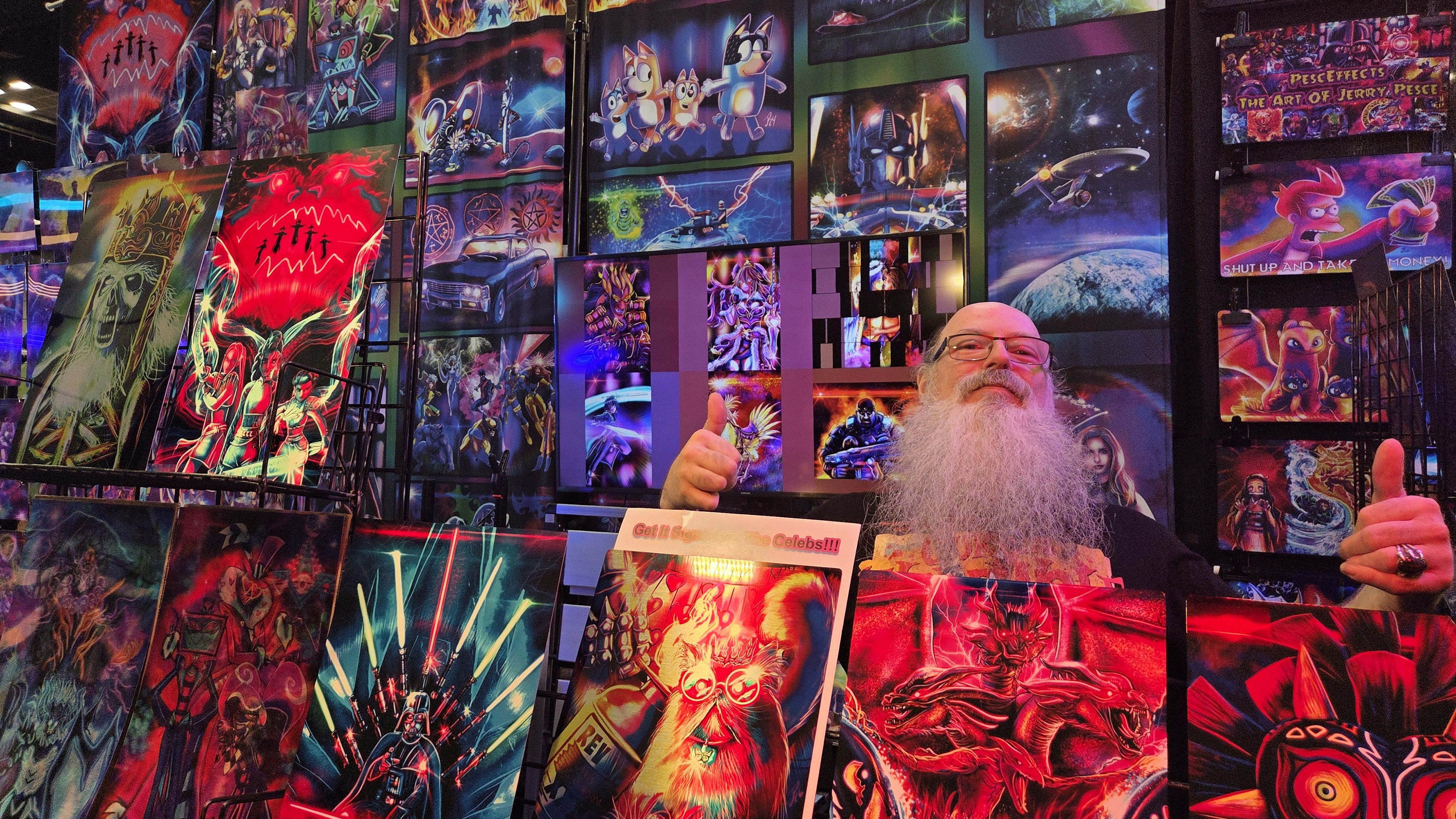 Bearded man with glasses behind a counter giving a thumbs-up, with neon-colored posters and canvases on the wall showing sci-fi, space, and cartoon artwork.