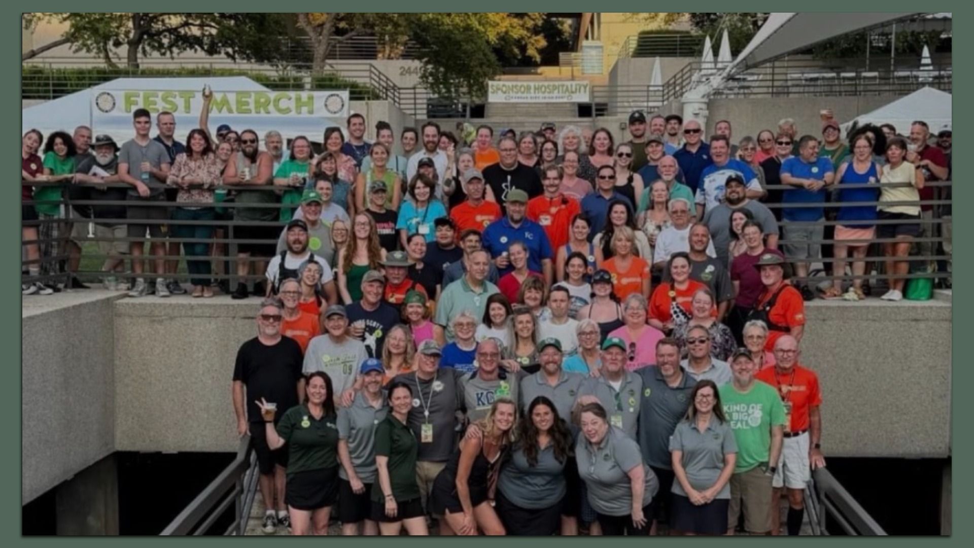 Large group photo at an outdoor festival, people posing on stairs with railings. Banners read "FEST MARCH" and "SPONSOR HOSPITALITY" in the background; crowd wears colorful shirts and smiles.