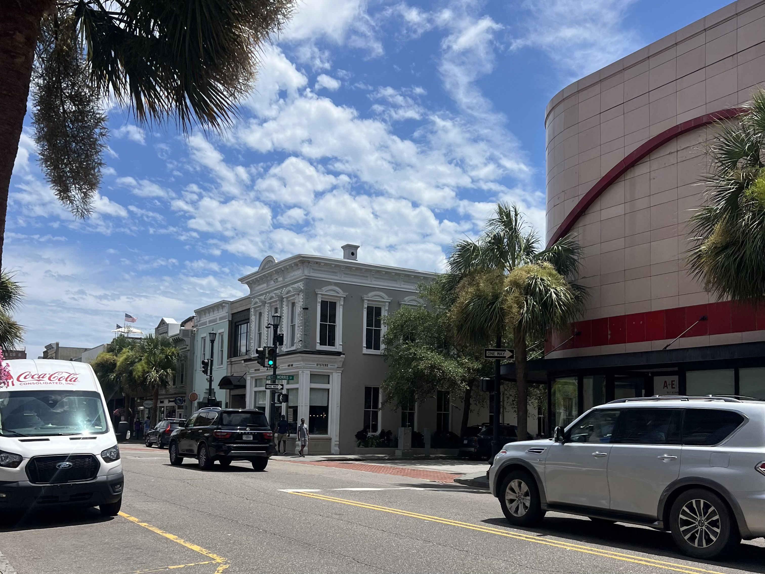 City street with palm trees and a blue sky. On the left a white ornate storefront, on the right a curved beige building with a red stripe. Front shows parked cars and a Coca-Cola van.