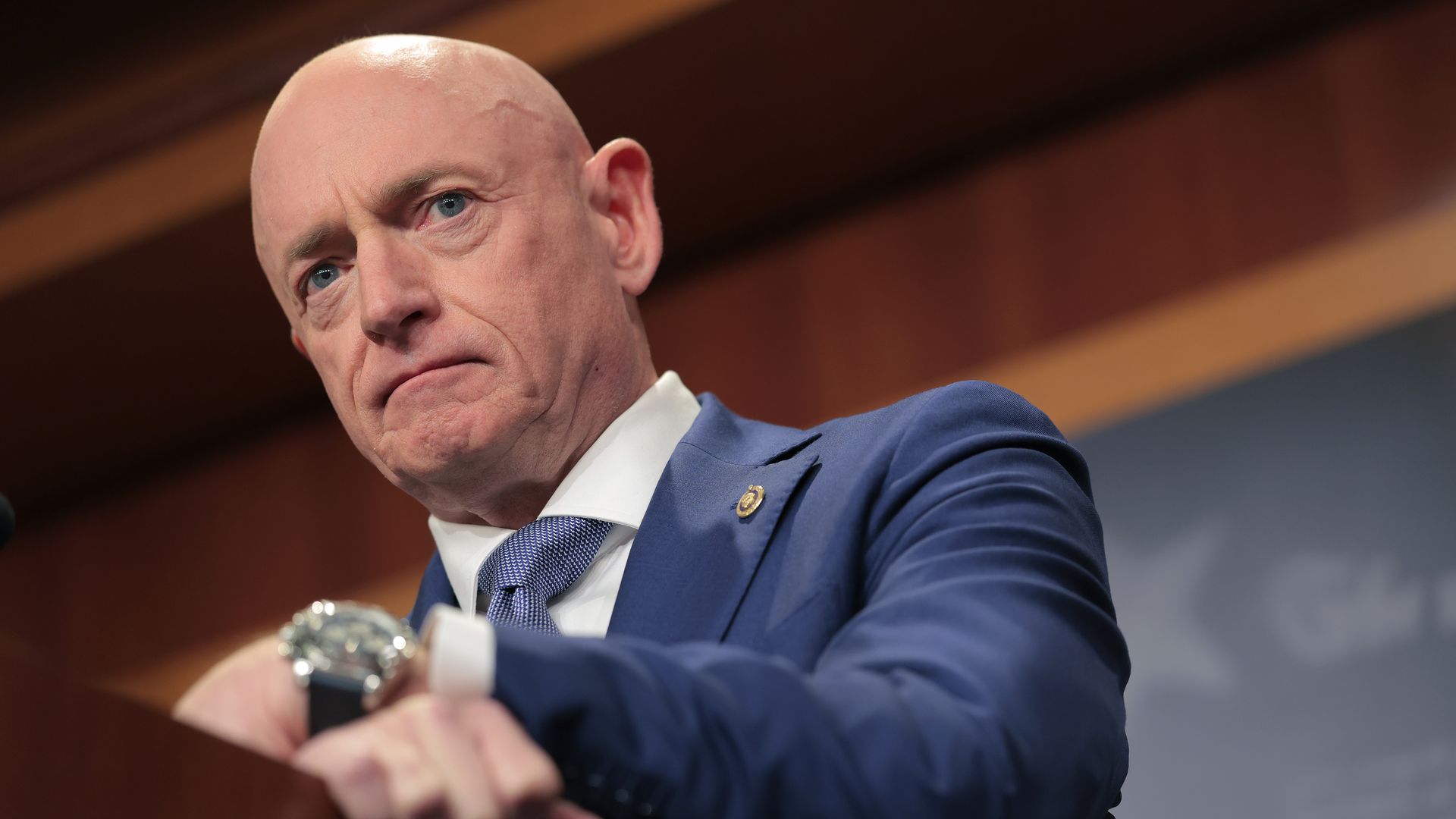 Bald man in a blue suit and tie standing behind a podium with a serious expression, light brown wooden background, and a blurred blue banner to the right.