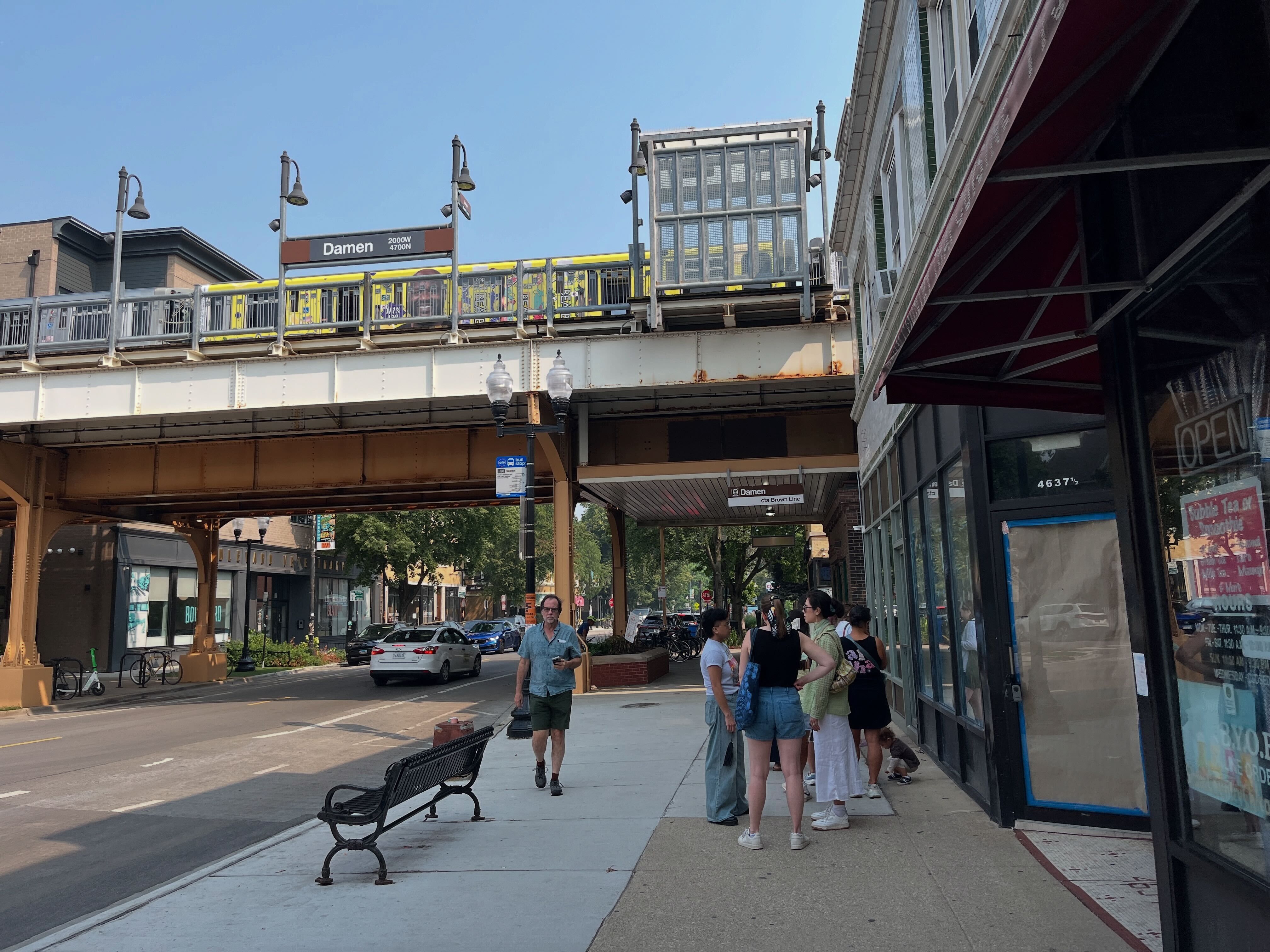 Street scene at Damen CTA Brown Line station with elevated train, people standing on sidewalk near storefronts, and a man walking by a bench under clear blue sky.