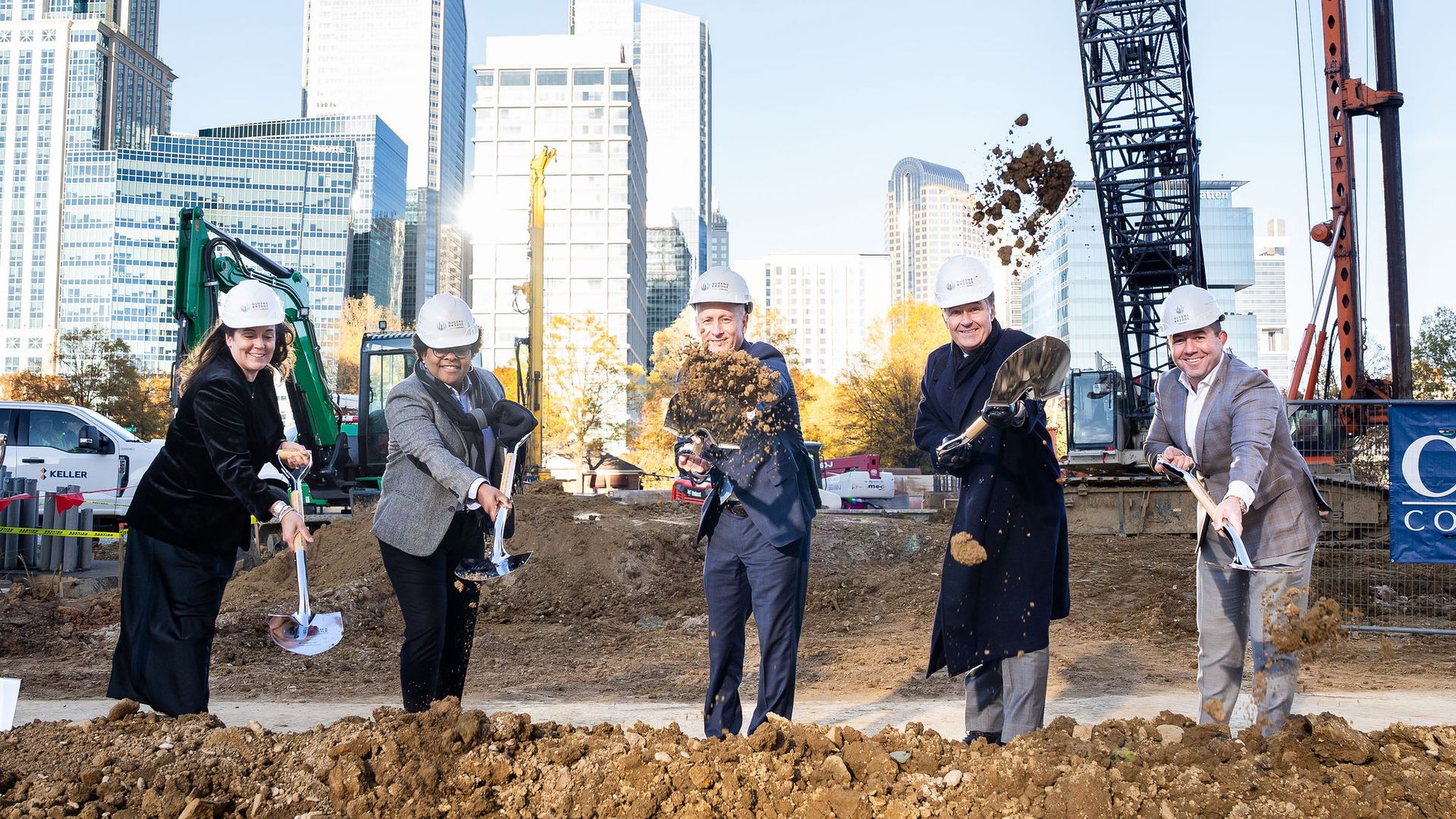 Five people in business attire and white hard hats participate in a groundbreaking ceremony, shoveling dirt at a construction site with urban skyscrapers in the background.