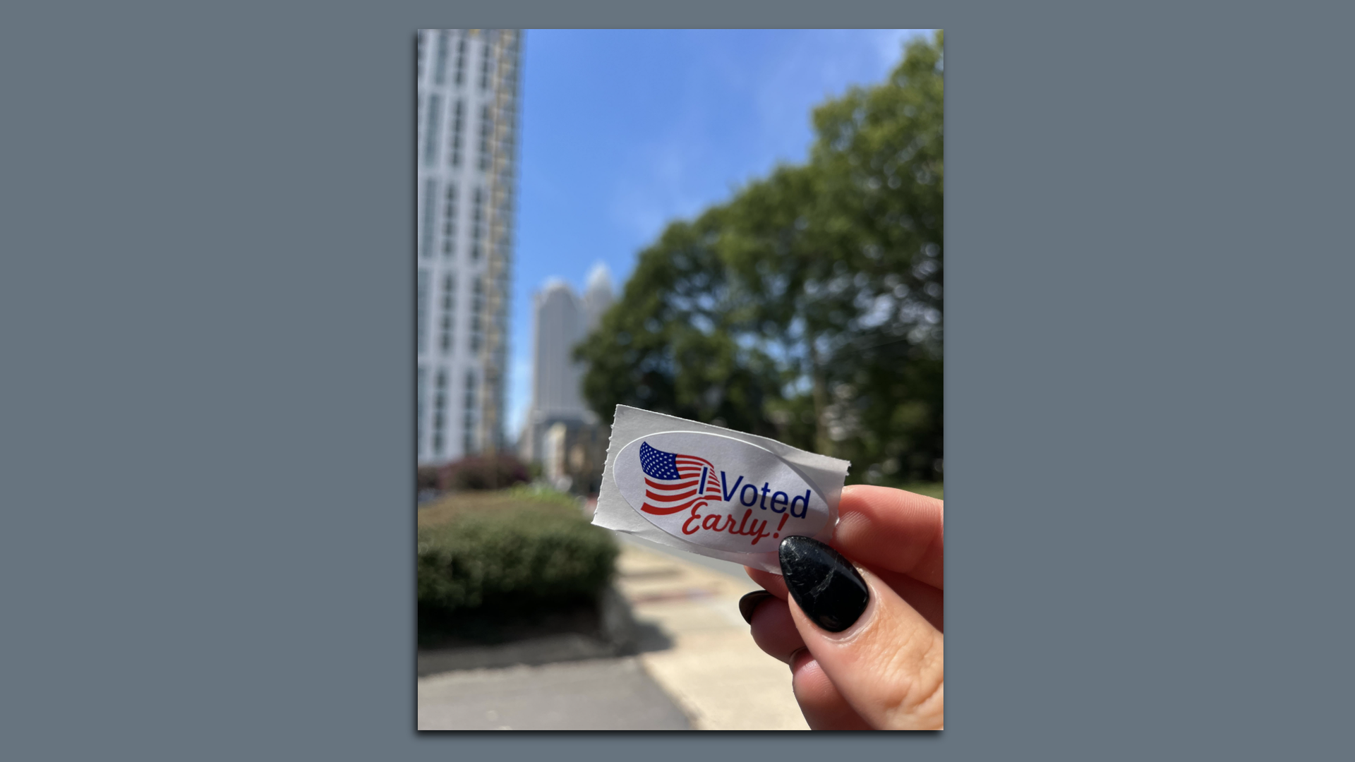Hand with black nail polish holding a "Voted Early!" sticker featuring an American flag, with a sunny outdoor city scene in the blurred background.