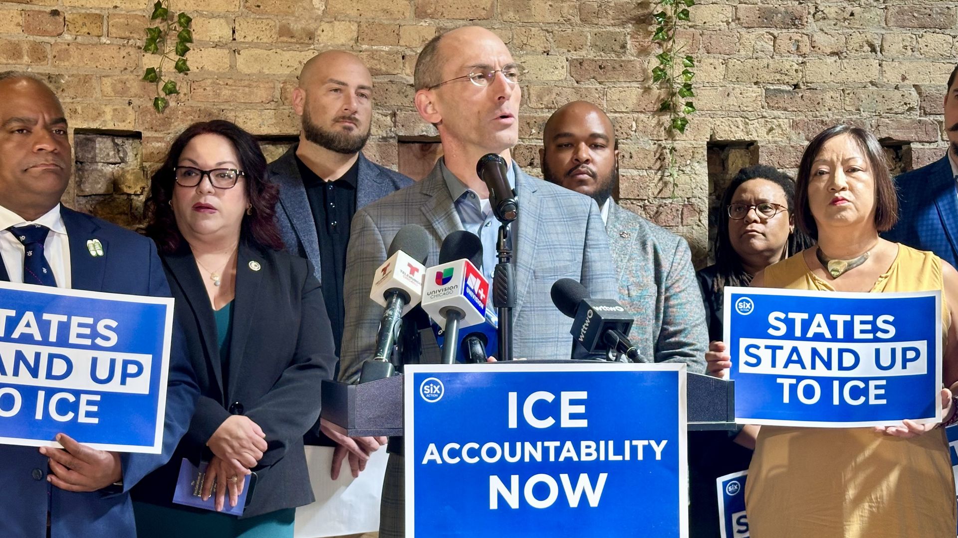 Diverse group at a press conference; central speaker in a gray suit with microphones, others hold blue posters reading "STATES STAND UP TO ICE" against a brick wall.