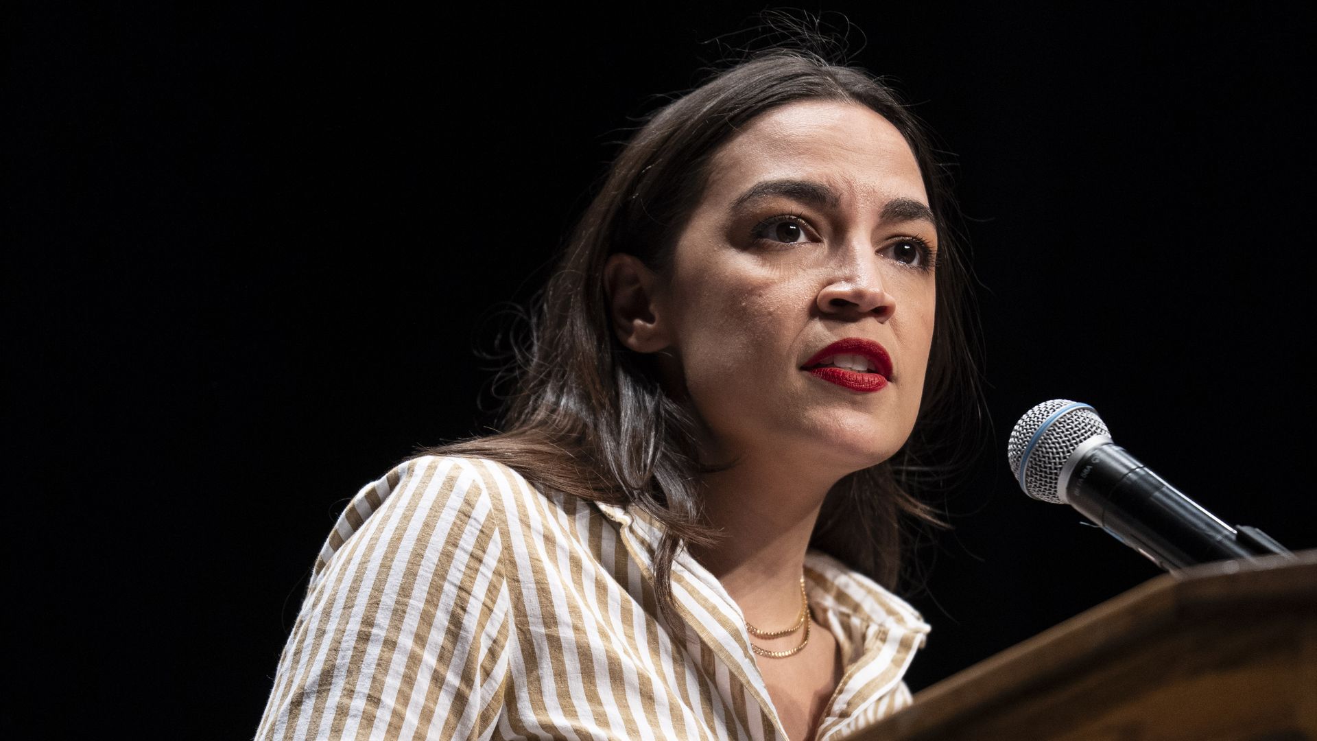 A woman with dark hair and red lipstick stands at a wooden podium, speaking into a microphone. She wears a beige and white striped blouse on a dark stage.