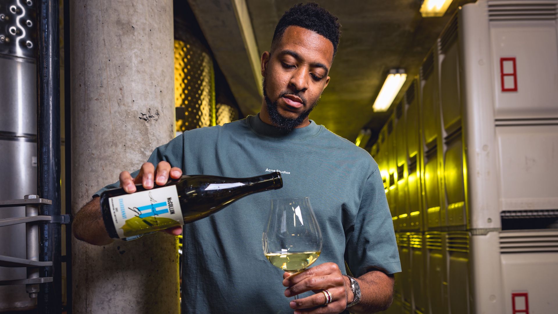 CJ McCollum, standing in an industrial looking space, pours a glass of white wine into a wine glass.