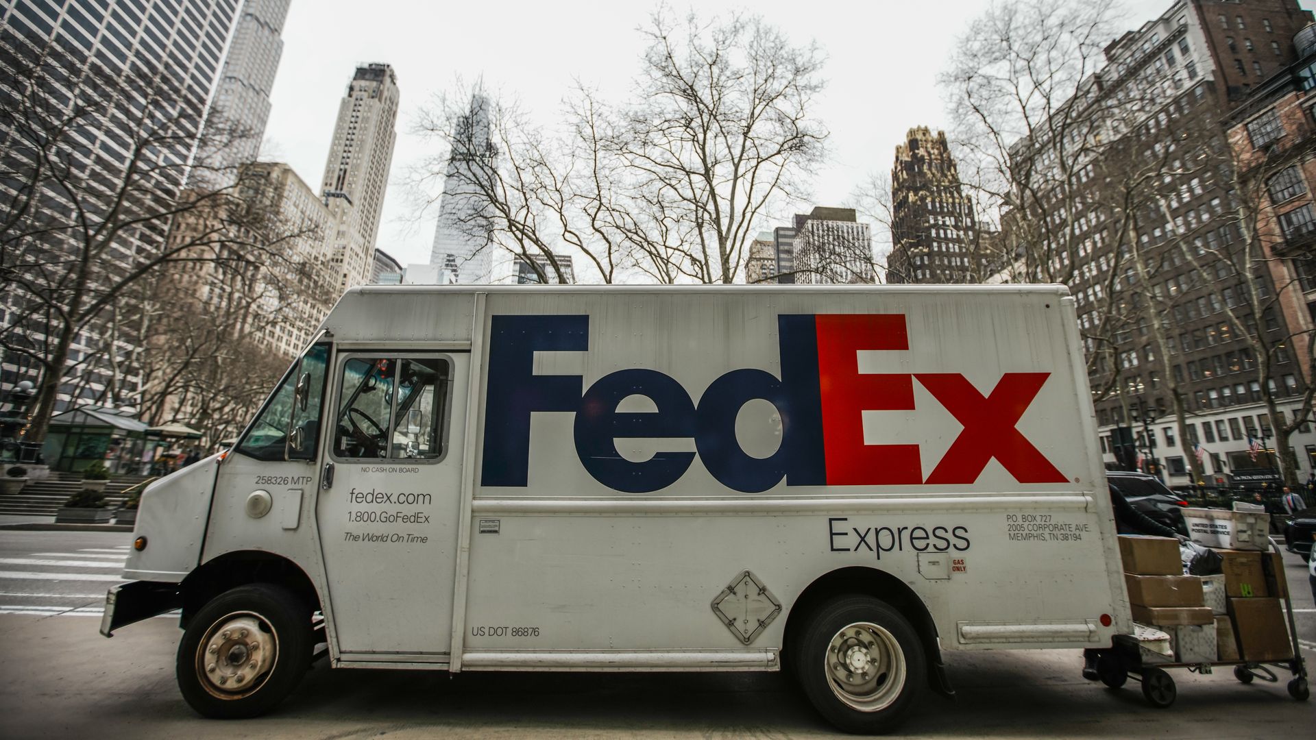White FedEx Express delivery truck on a city street, with a large blue Fed and red Ex logo on its side; bare trees and tall skyscrapers loom in the background.
