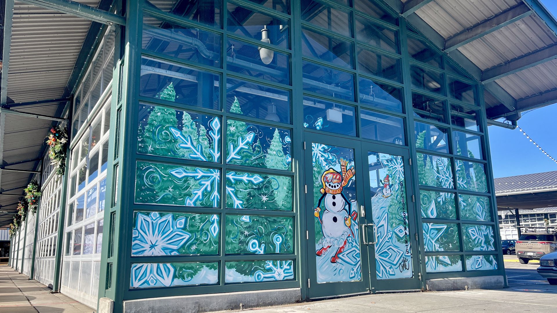 Green-framed glass building corner decorated with festive paintings of large blue snowflakes, green pine trees, and a snowman wearing an orange scarf and red skis on a snowy background.
