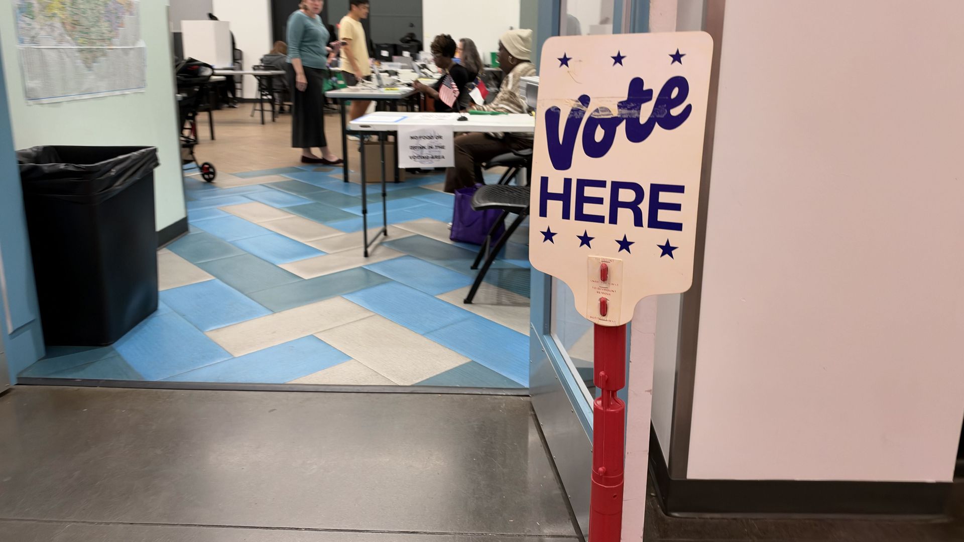 Inside a polling station, a large white "Vote Here" sign with blue letters and stars stands in the foreground. People sit at tables with forms; a map on the left and blue tiled floor.