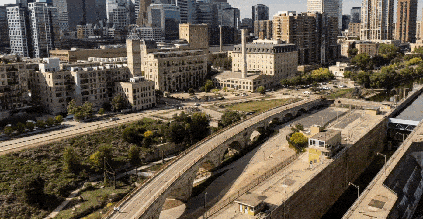 Aerial view of a historic stone arch bridge over a river lock, surrounded by city buildings with green trees and skyscrapers in the background under a clear sky.