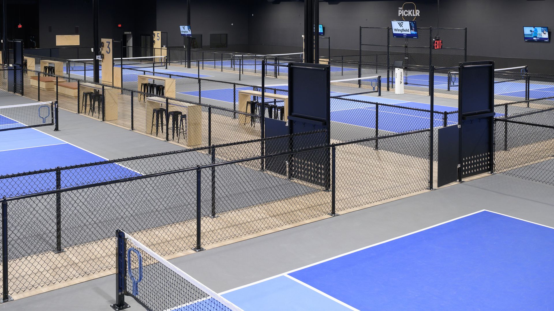 Indoor pickleball courts with blue and gray floors, black chain-link fences, wooden seating areas, digital scoreboards, and dark walls under bright ceiling lights.