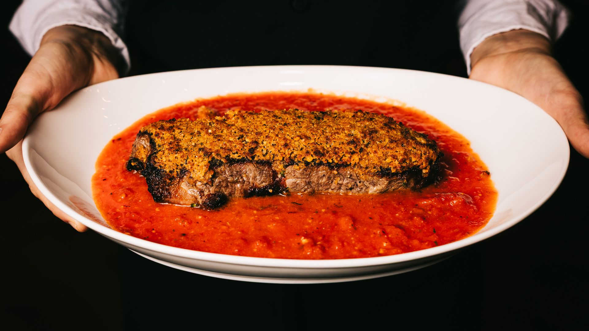 Person holding a white plate with a crusted steak served on a pool of chunky red tomato sauce, against a dark background.