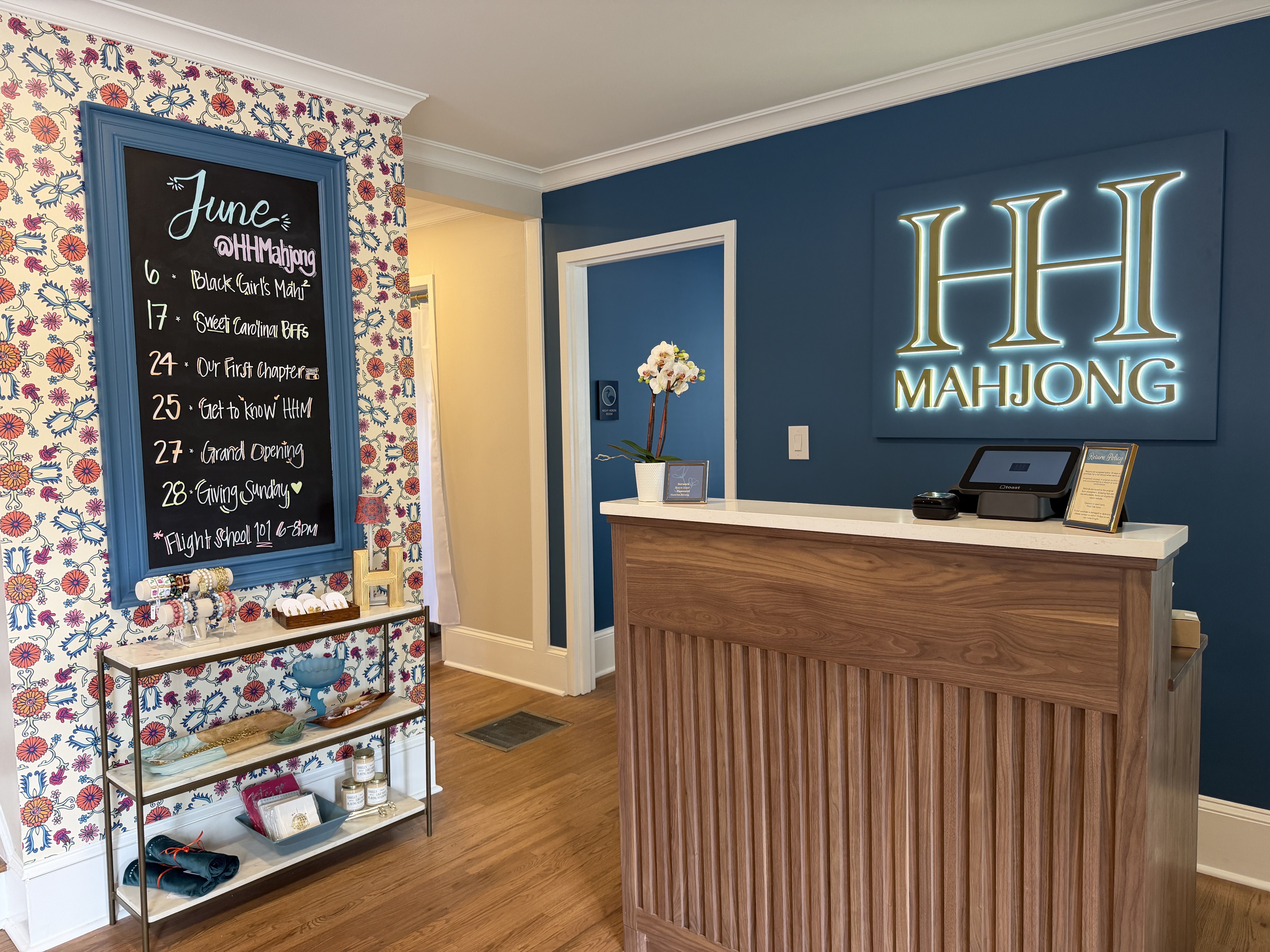 Interior of a boutique with a blue wall and neon "HH MAHJONG" sign. A teal-framed chalkboard lists June events beside floral wallpaper; a wooden reception desk with a plant and product display.